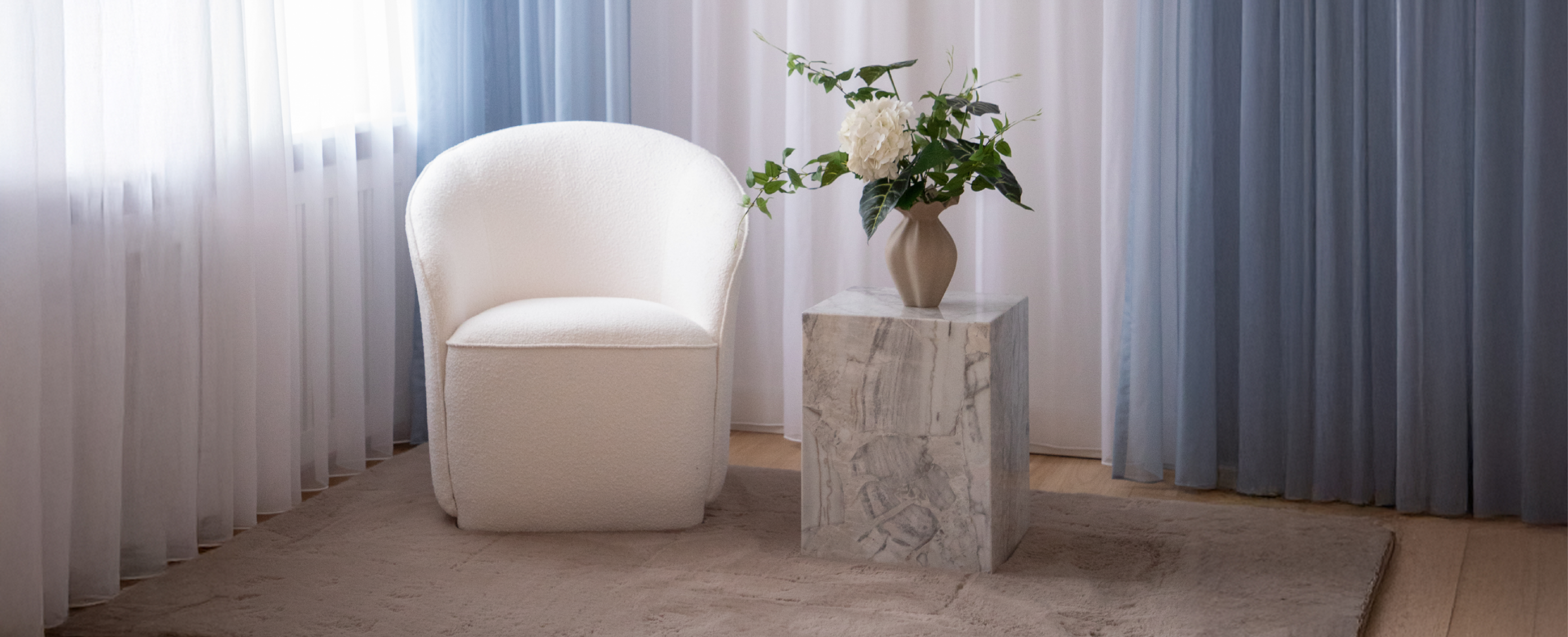 Hvid loungestol fra Specktrum. Ved siden af står et marmor sidebord, med en brun keramik vase og kunstige blomster. På gulvet ligger et beige gulvtæppe. Baggrunden er gulv til loft gardiner.