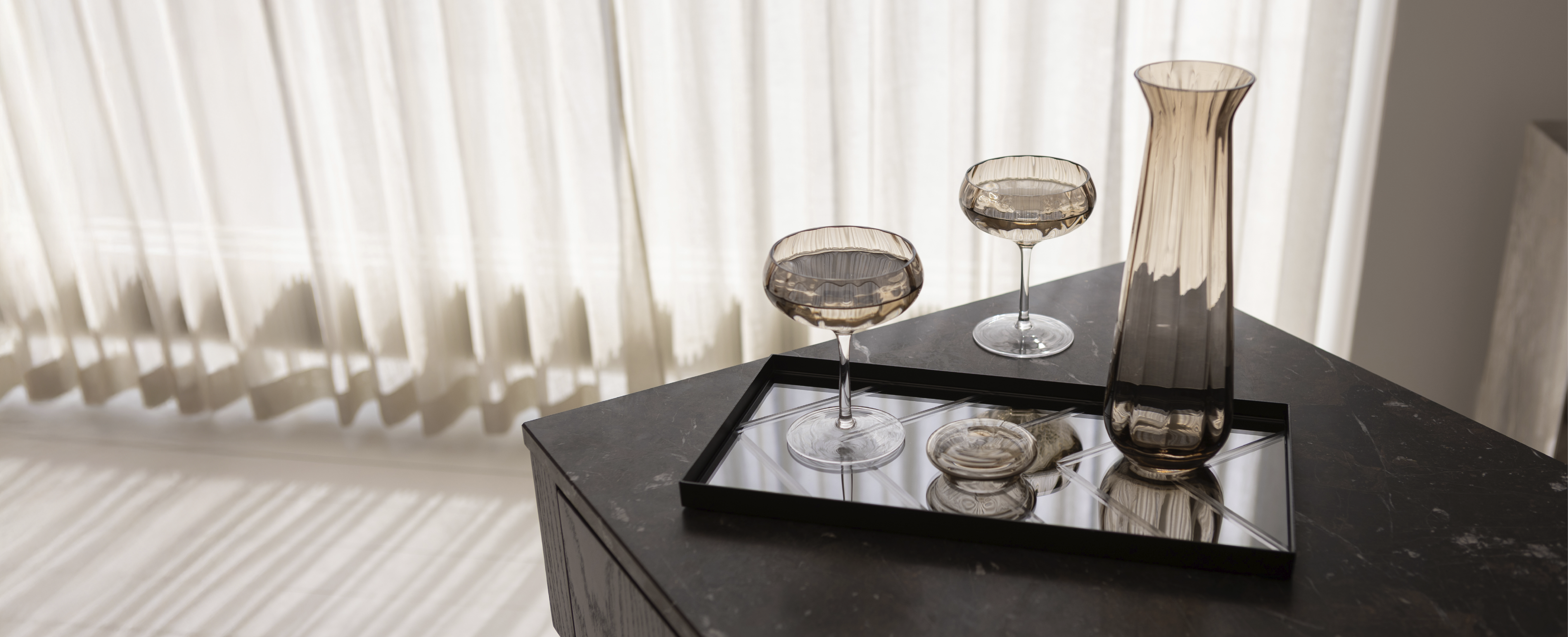 En spejlbakke med sort metal ramme, der står på et mørkt køkkenbord. På bakken står en glas karaffel og to cocktail glas i brun. Baggrunden er hvide gardiner.