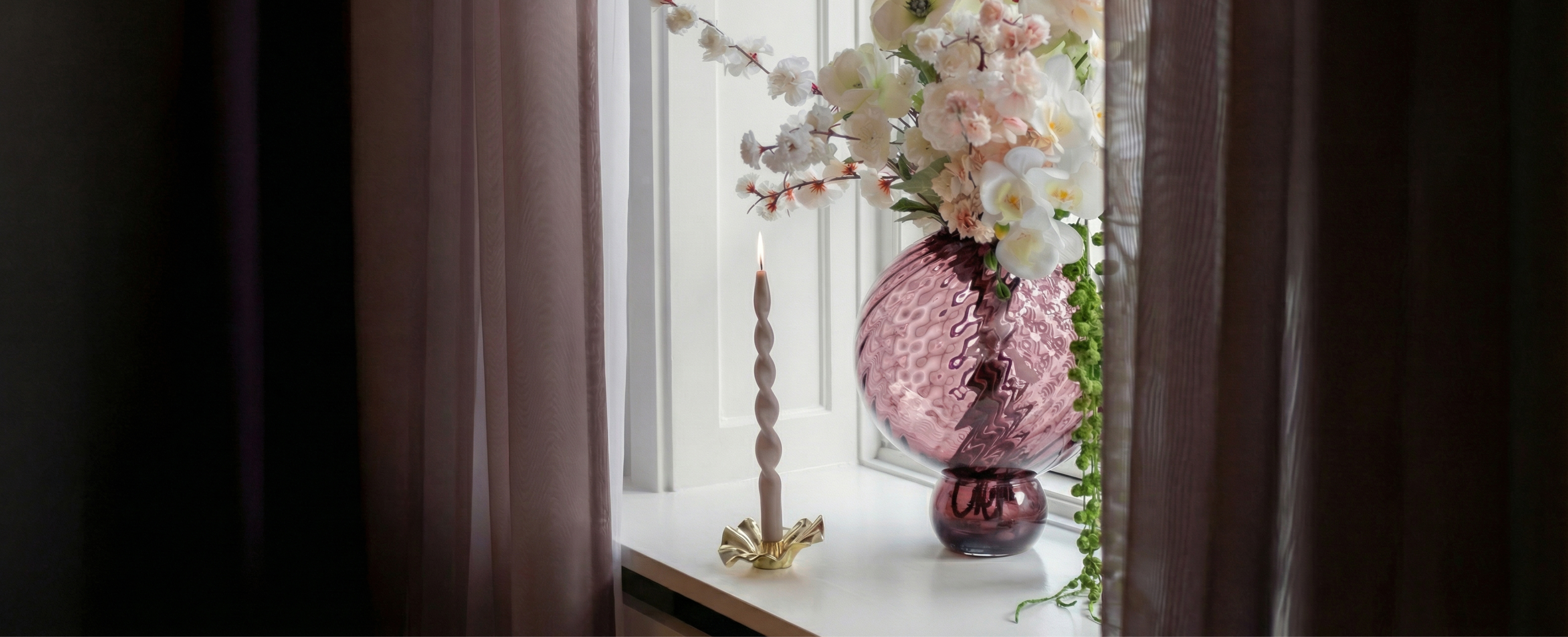 Lyserød glas vase med en buket kunstige blomster, der står i en vindueskarm. Ved siden af står en organisk guld metal lysestage med et tændt snoede lys. I begge sider af vinduet hænger mørke rosa gardiner.