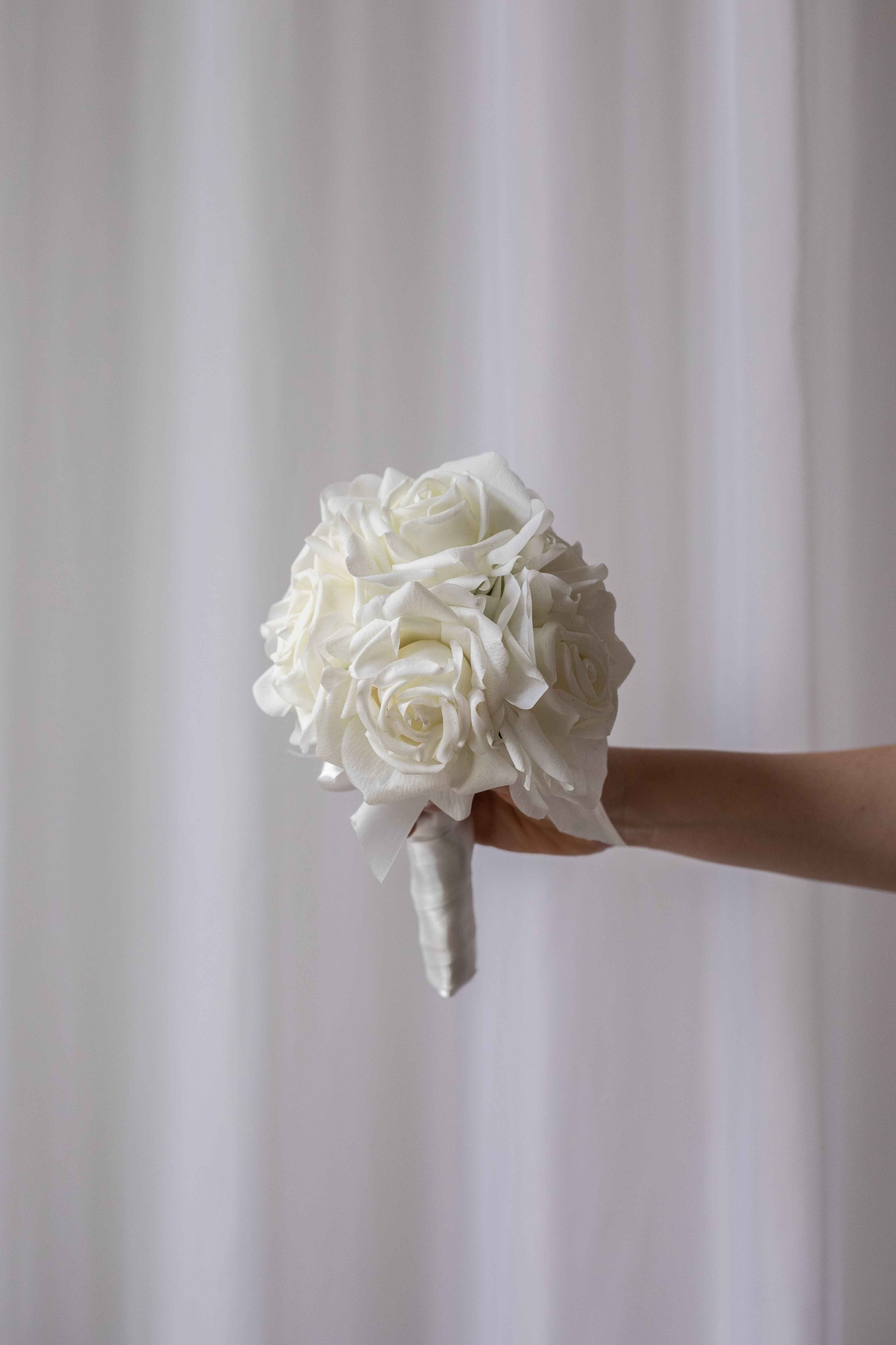 Brudebuket 5 - bestående af 7 hvide roser. Buketten holdes op foran et hvidt gardin.