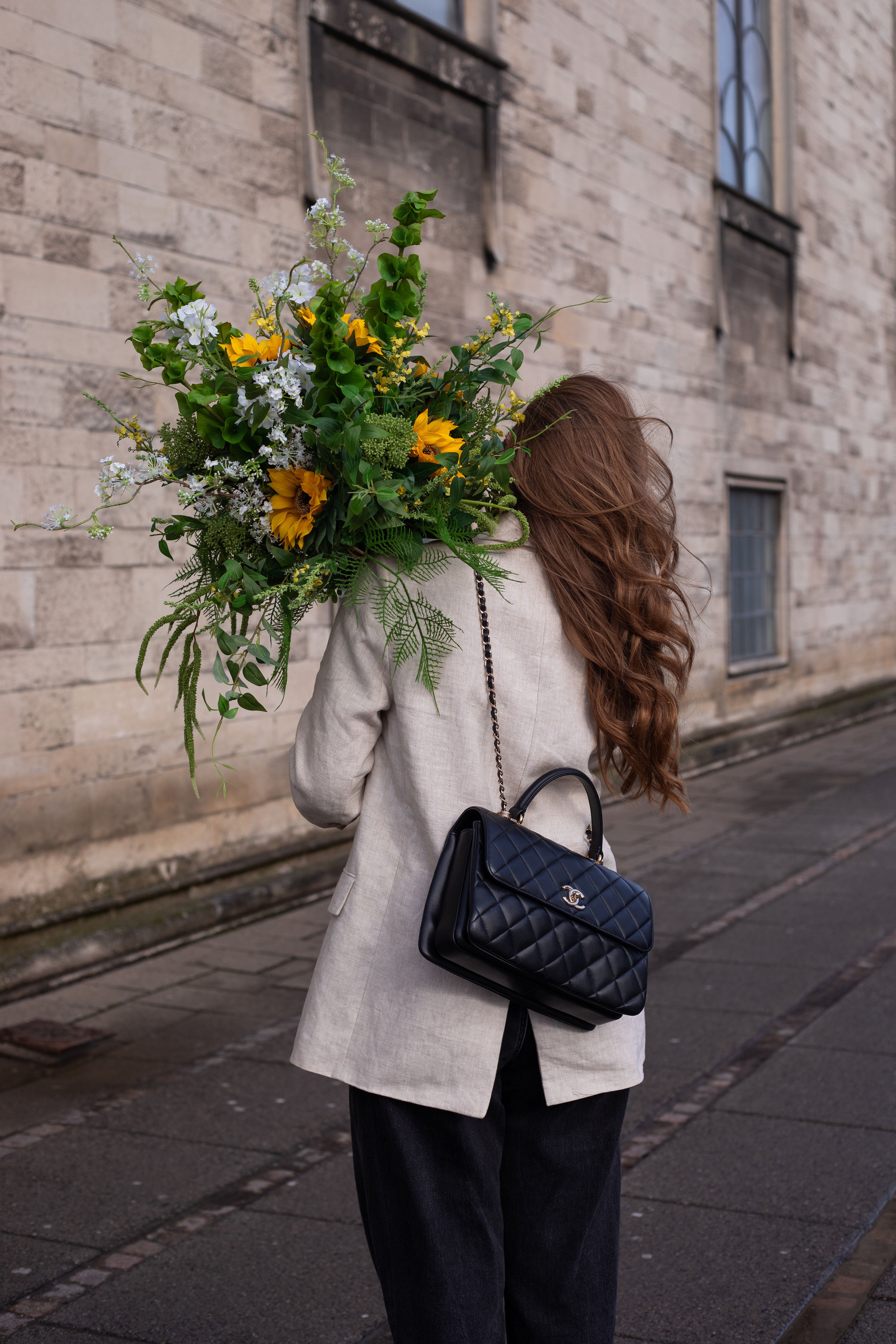 Pige med langt brunt hår, beige blazer og sort Chanel taske, der holder en stor kunstig buket i hvide, grønne og gul nuancer over skulderen. Billedet er taget udenfor, foran en beige murstens bygning.