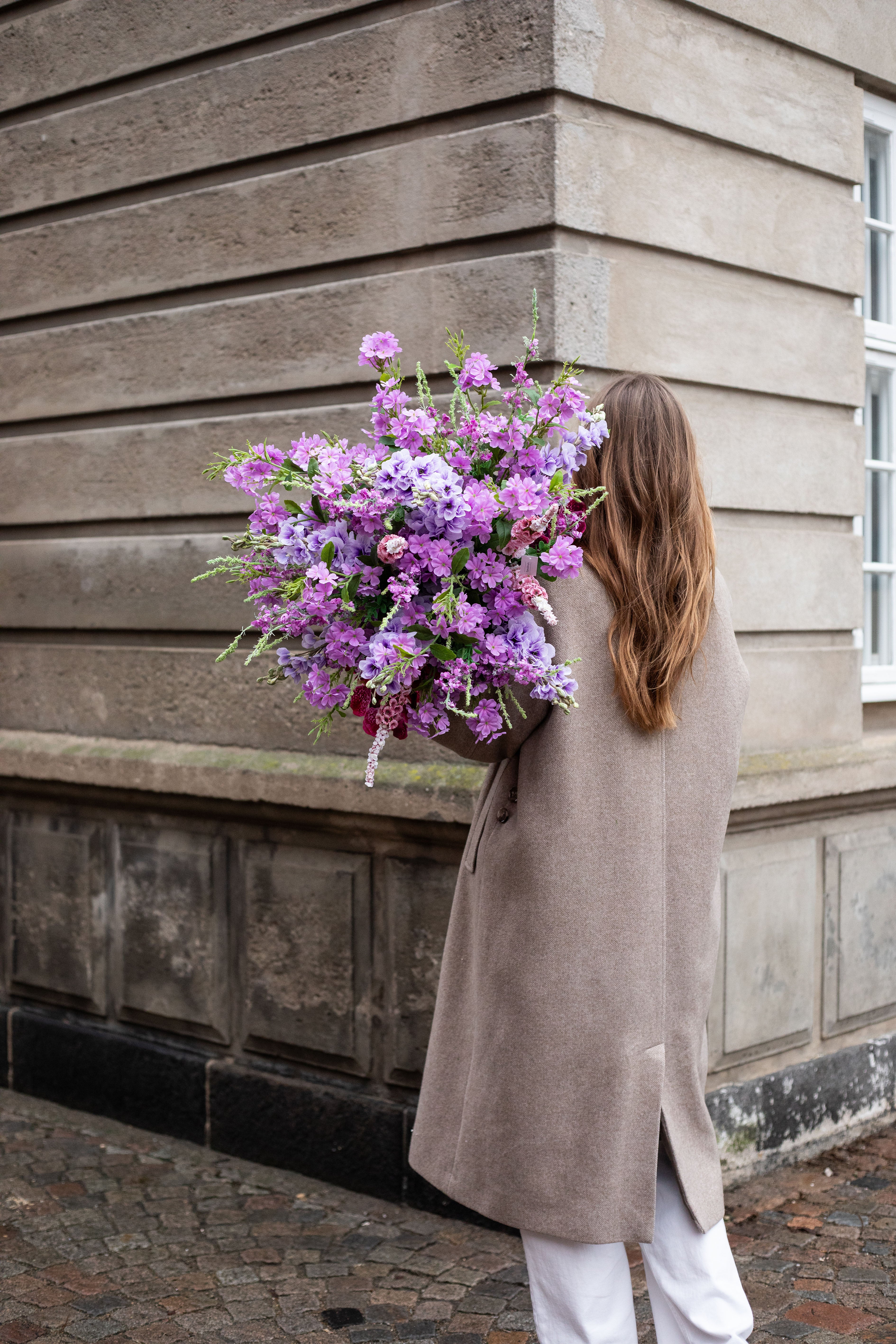 En pige står med ryggen til, hun har langt brunt hår, beige frakke og hvide jeans på. Over skulderen holder hun en stor kunstig buket i lilla og rosa nuancer. Billedet er taget udenfor foran en beige bygning.