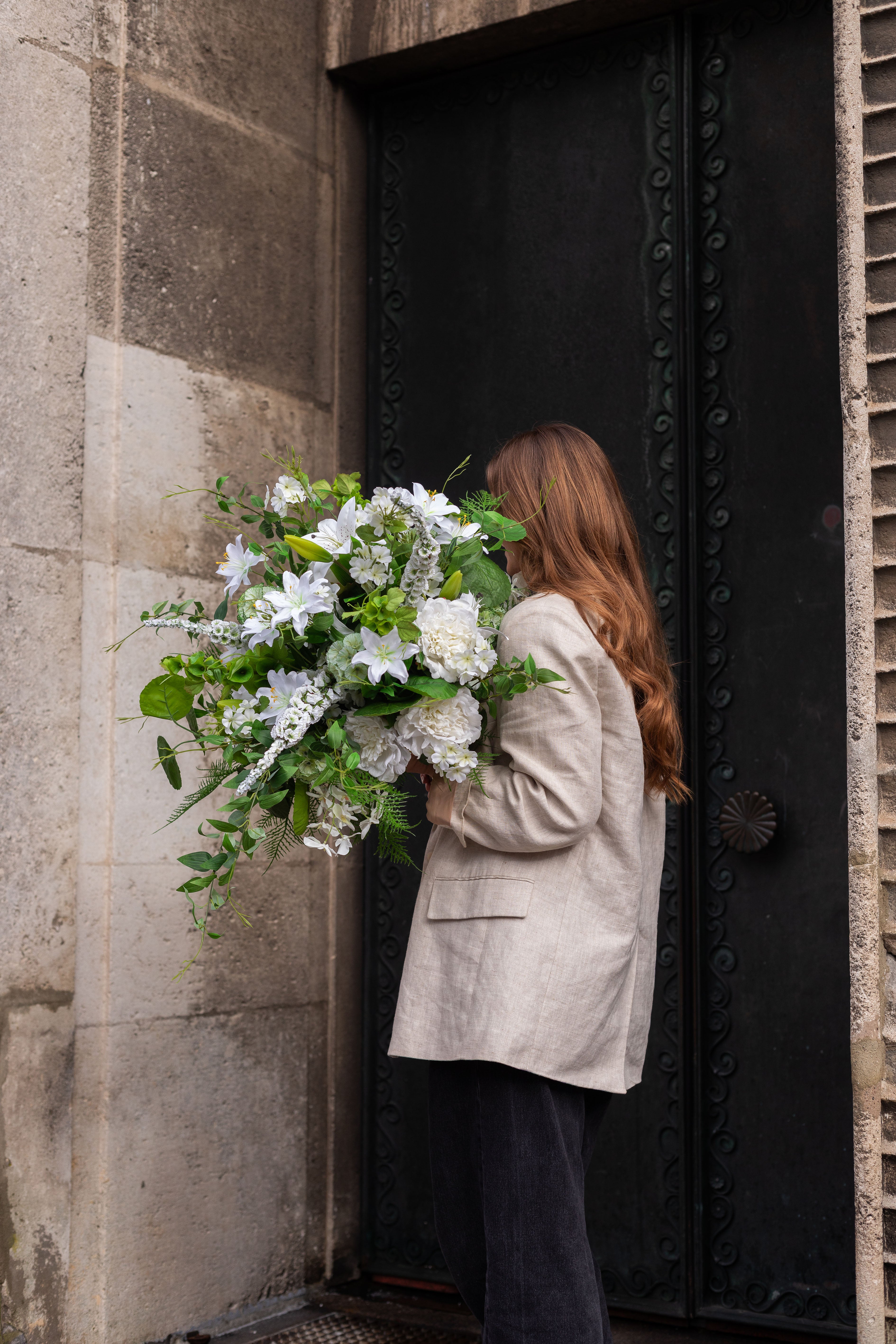 Pige der står med ryggen til. Hun har langt brunt hår, beige blazer og sorte jeans. I hånden holder hun en stor kunstig buket i grønne og hvide nuancer. Billedet er taget foran en beige bygning, med en stor sort metal dør.