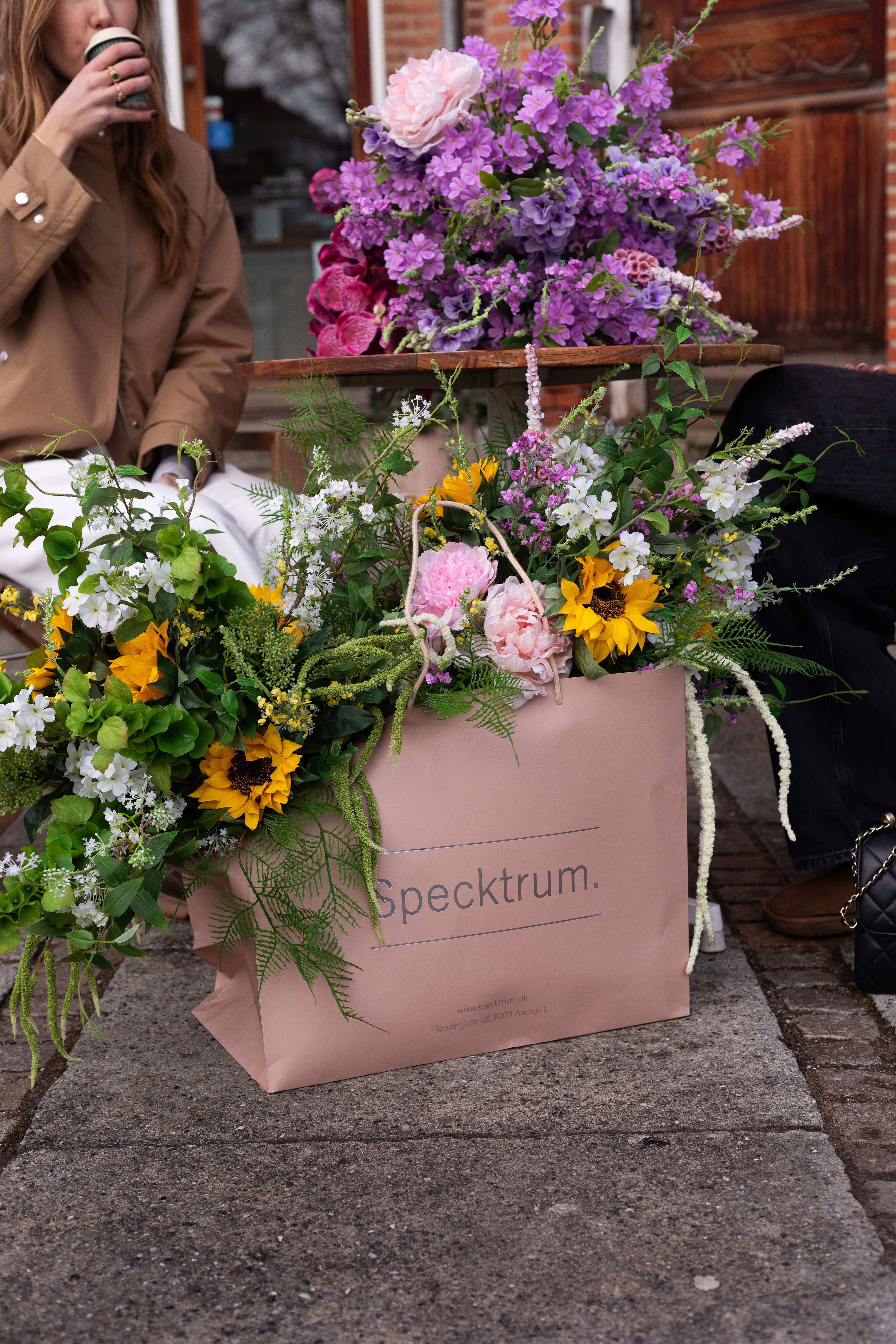 Billedet af lyserød Specktrum pose, med kunstige buketter i, der står på asfalt. Bagved sidder to piger og drikker kaffe ved et bord, på bordet ligger en stor kunstig buket.