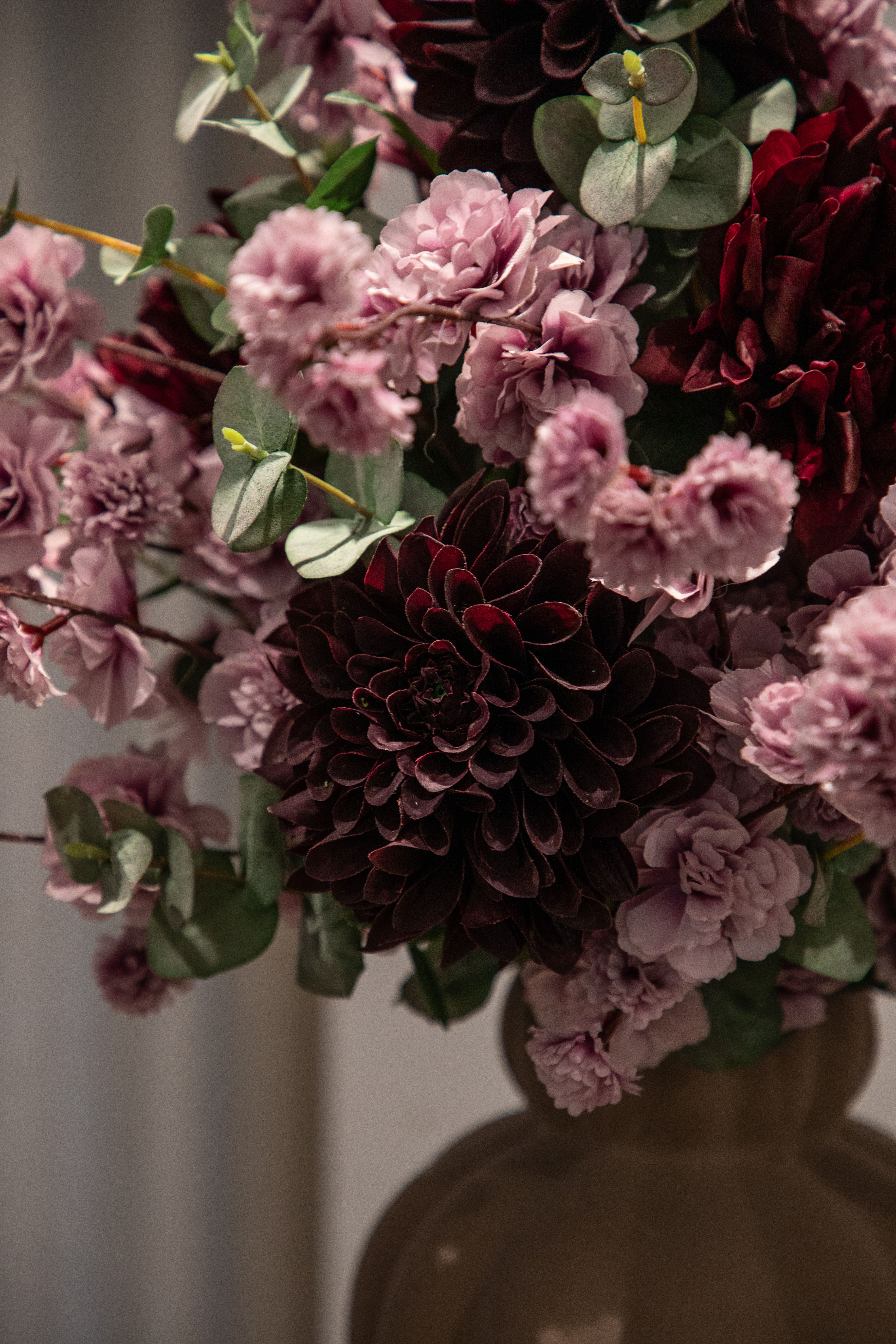 Nærbillede af stor kunstig buket i grønne, rose og bordeaux nuancer, der er placeret i en brun keramik vase.