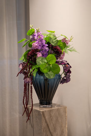 Stor kunstige buket i grønne, lilla og mørkerøde nuancer, placeret i en blå glas vase, der står på en marmor piedestal.