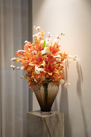 Stor størrelse kunstig buket i hvide og orange nuancer, der er placeret i en røgfarvet glas vase, der står på en marmor piedestal.