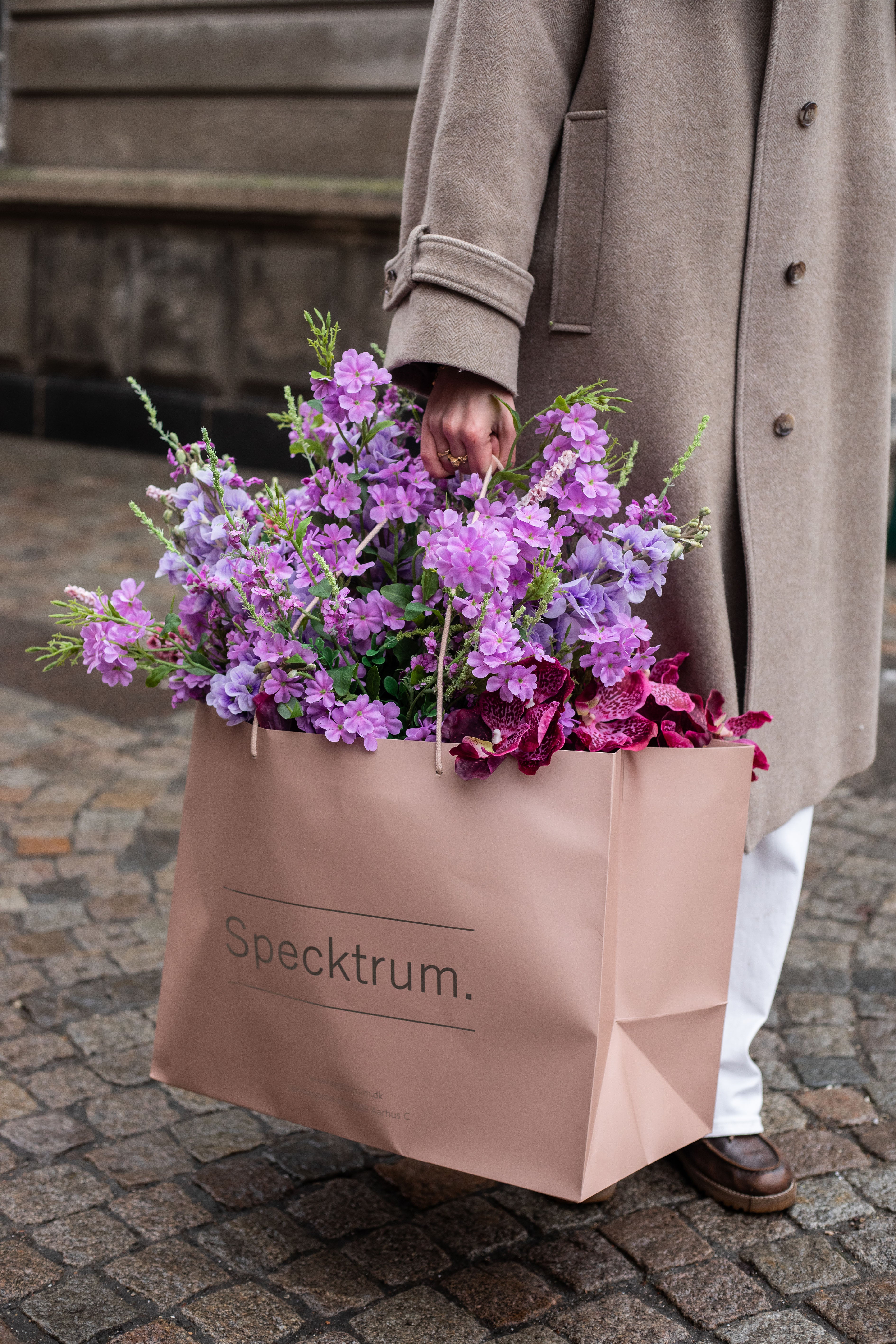 En pige i beige frakke og hvide bukser, der holder en lyserød Specktrum pose med kunstig buket i lilla og rosa nuancer i hånden.