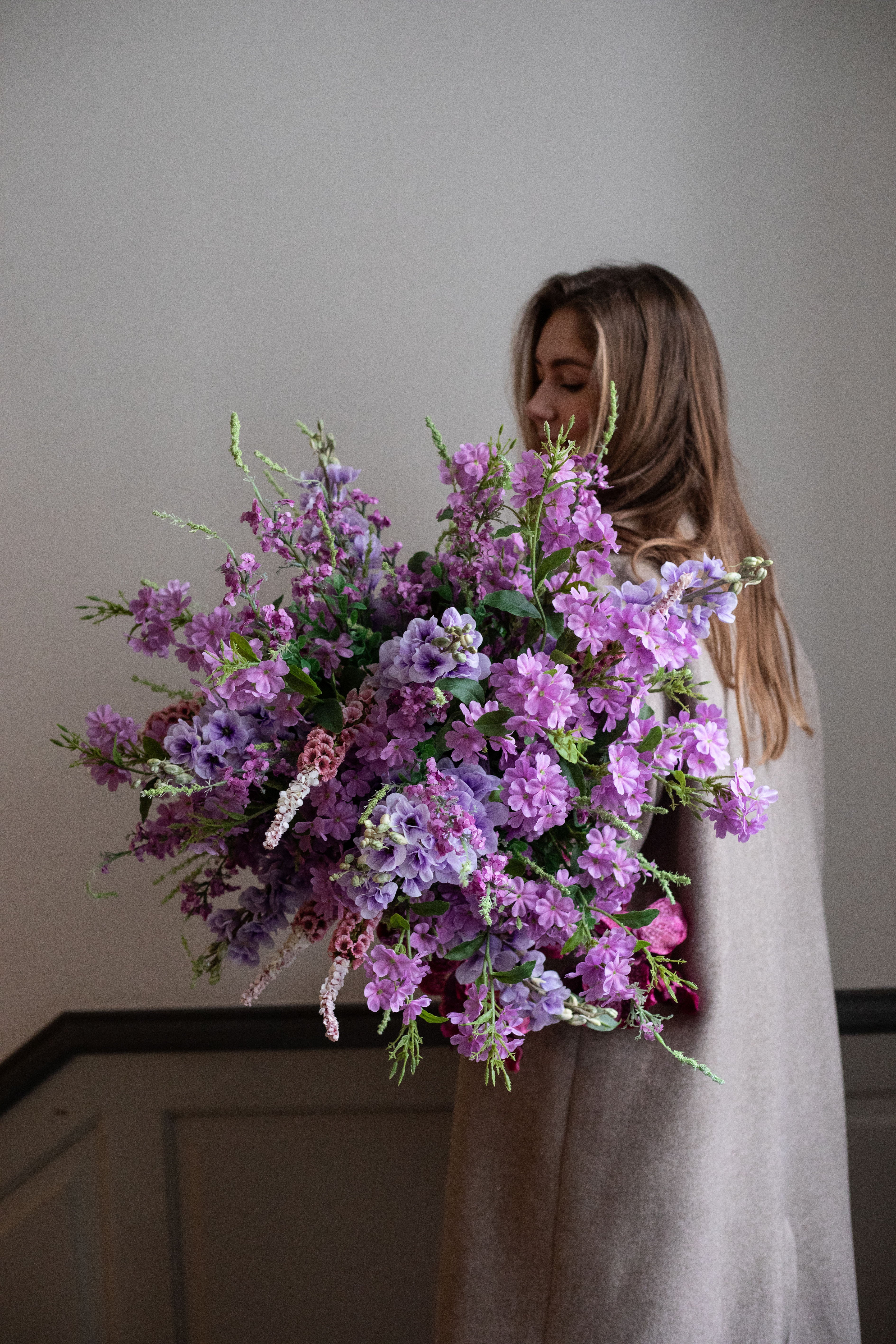 Pige med langt brunt hår og beige frakke, der holder en stor kunstig buket i rose og lilla nuancer fra Specktrum. Billedet er taget i en opgang.