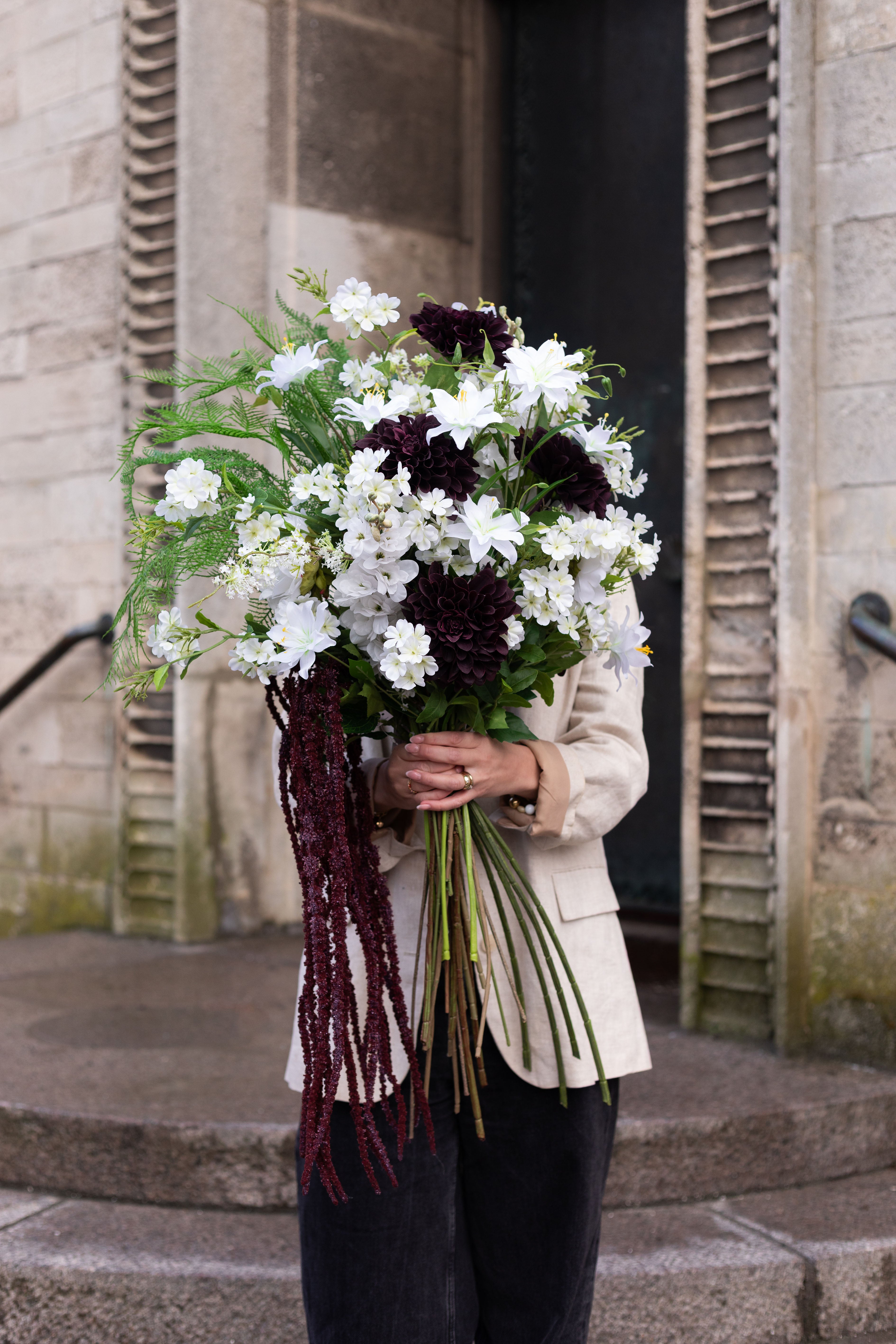 Pige der holder en stor kunstig buket i hvide, grønne og bordeaux nuancer op foran hovedet. Hun har en beige blazer og sorte jeans på. Billedet er taget på en trappe foran en beige murstens bygning.