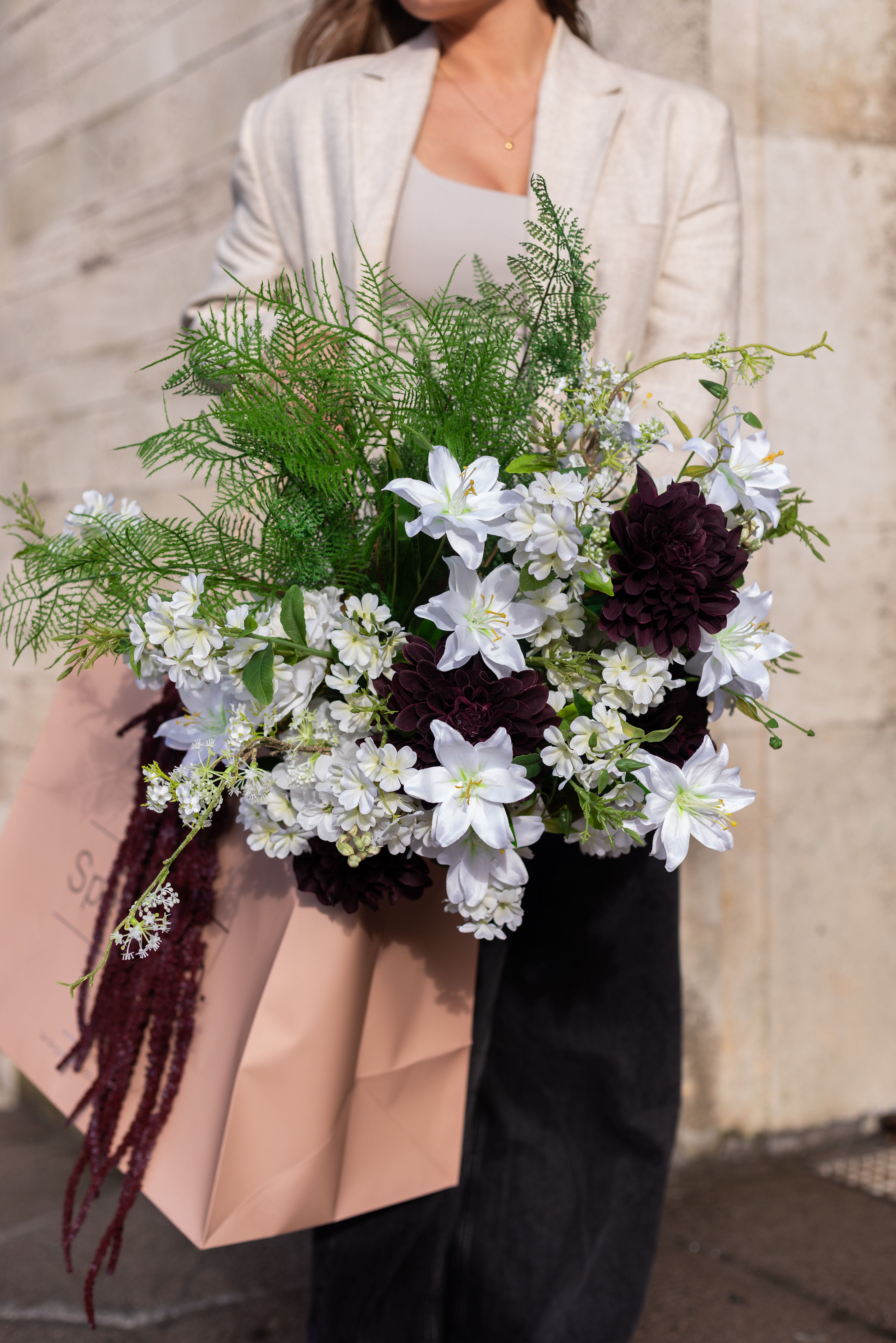 Pige i beige blazer og sorte jeans, der holder en lyserød Specktrum pose, hvori der er kunstige blomster i hvide, grønne og bordeaux nuancer.