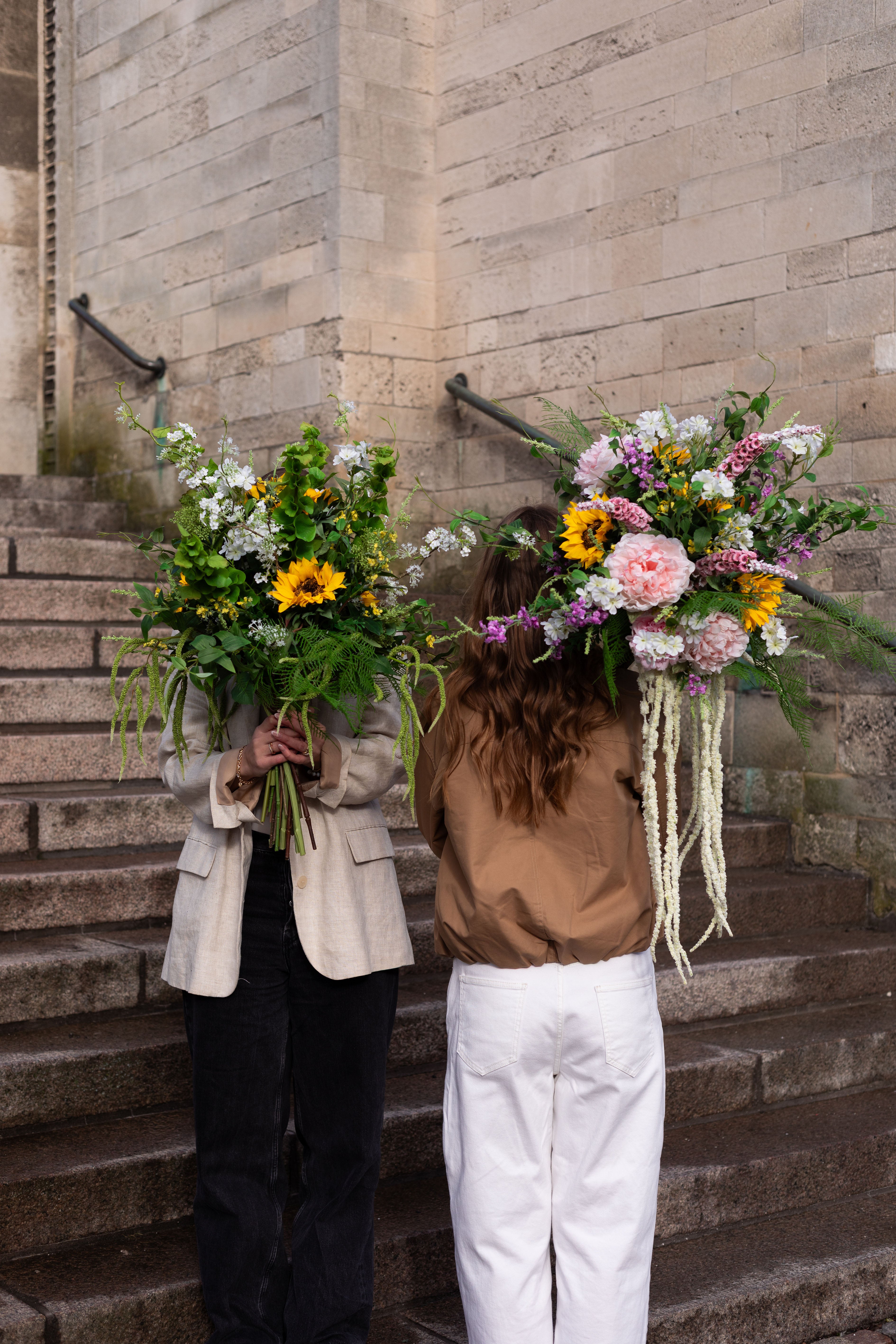 To piger, der står på en trappe foran en beige bygning. Pigen til højre holder en kunstig buket foran hovedet i grønne, hvide og gule nuancer. Pige til venstre holder en kunstig buket i rose, gule, hvide og lilla nuancer over skulderen med ryggen til kameraet.
