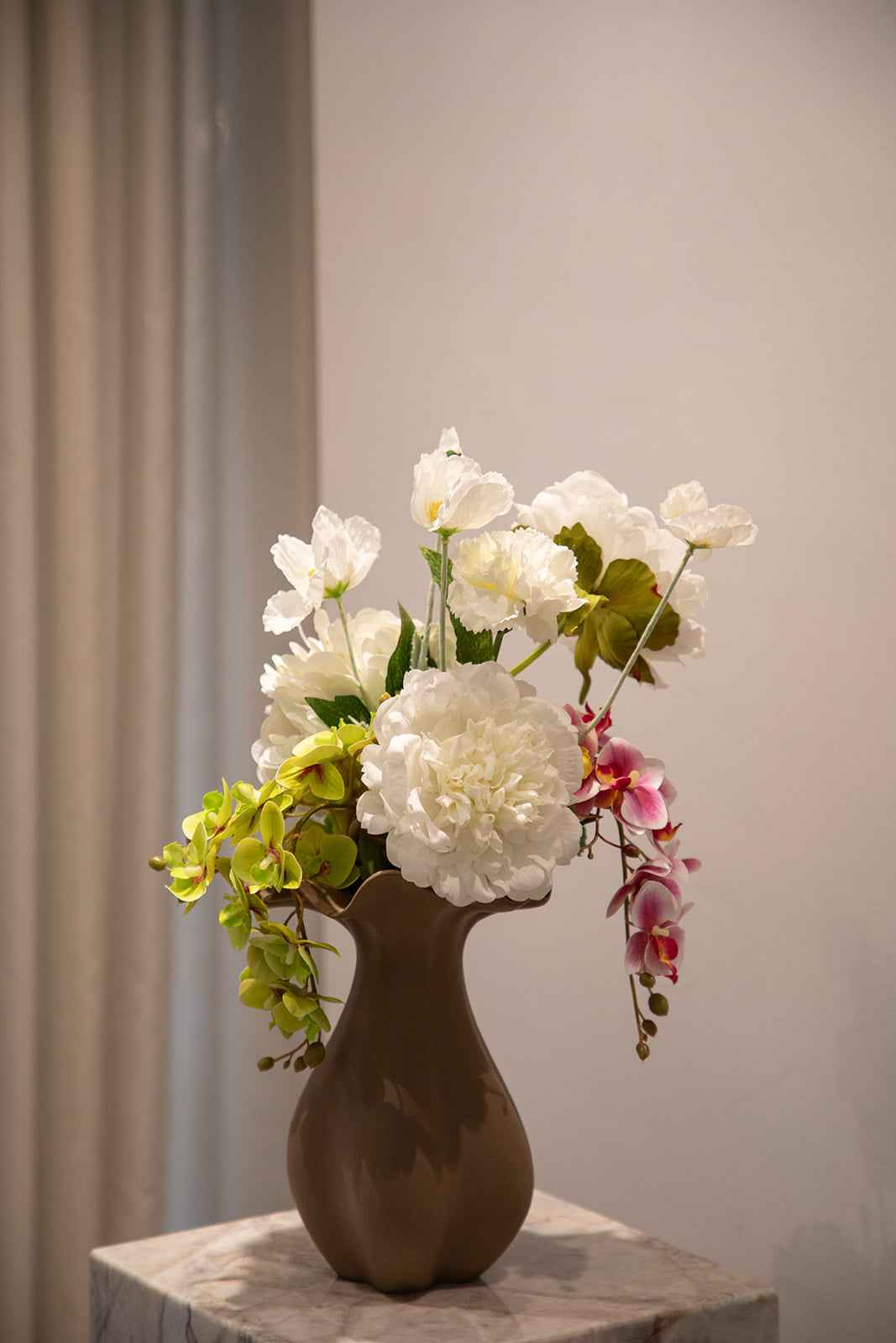 Buket af kunstige blomster i hvide, grønne og rosa nuancer. Placeret i en brun keramikvase, der står oven på en marmorpedestal.