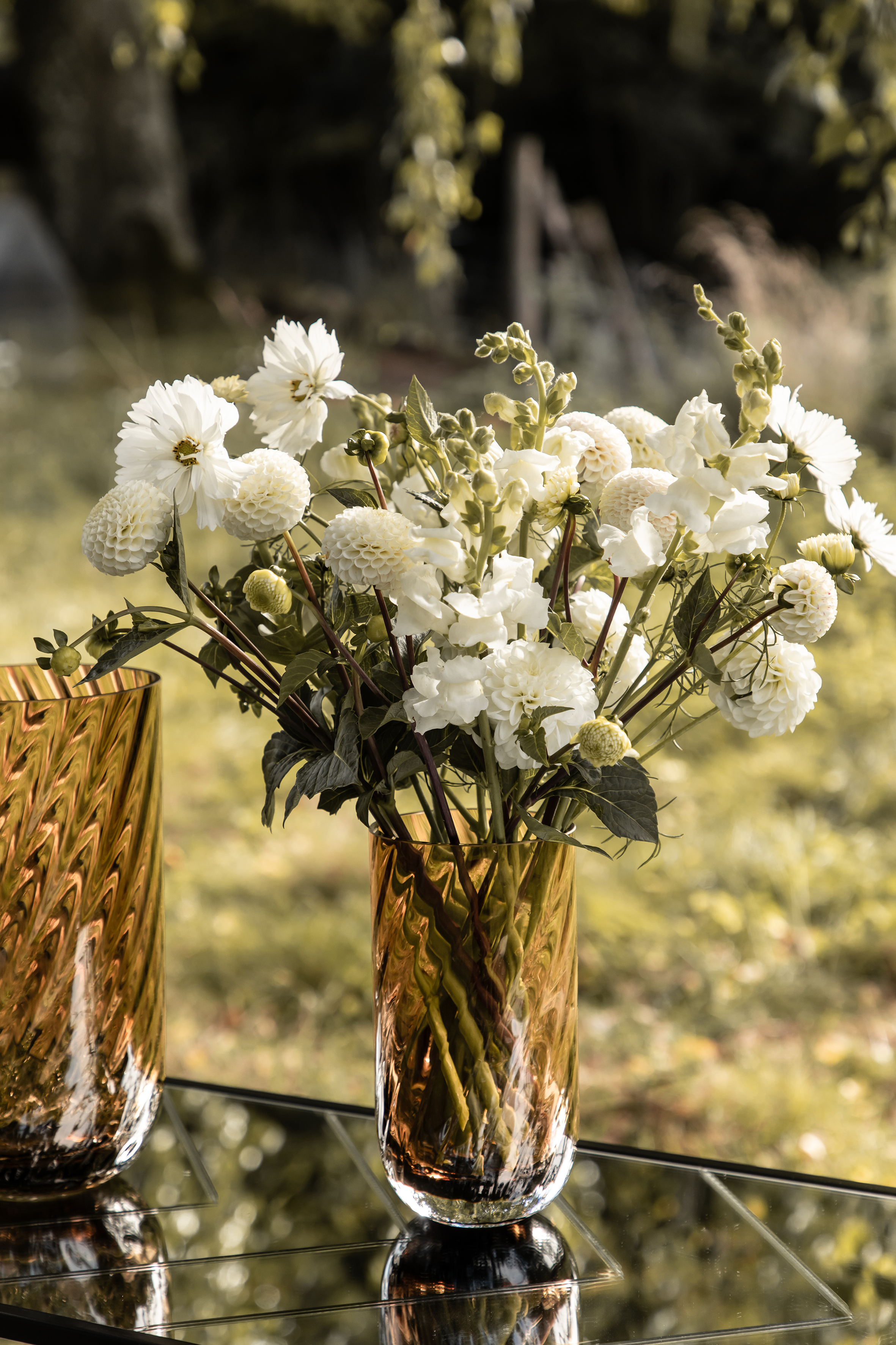 Cylindrisk ravfarvet glasvase med mønster fyldt med hvide blomster placeret på et spejlglasbord udendørs. En anden tilsvarende vase ses delvist i billedet.