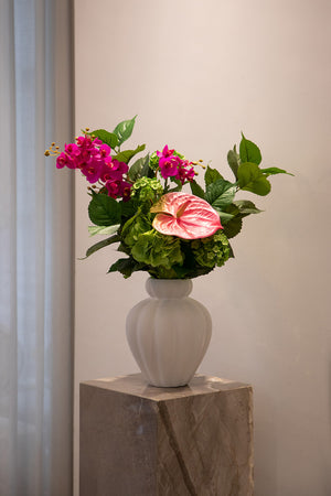 Billede af en basis buket af kunstige blomster i grønne nuancer, der er tilføjet få lyserøde stilke. Buketten er placeret i en hvid keramik vase, der står på en marmor piedestal.