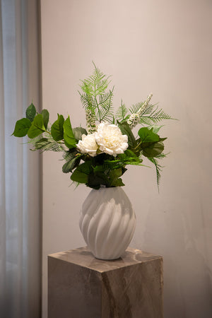 Kunstig blomster i hvide og grønne nuancer, der står i en hvid keramik vase, placeret på en marmor piedestal.