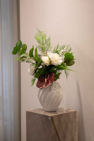 Hvid og grøn basis buket af kunstige blomster, hvor der er tilføjet en burgundy stilk for kontrast. Buketten er placeret i en hvid keramik vase, der står på en marmor piedestal.