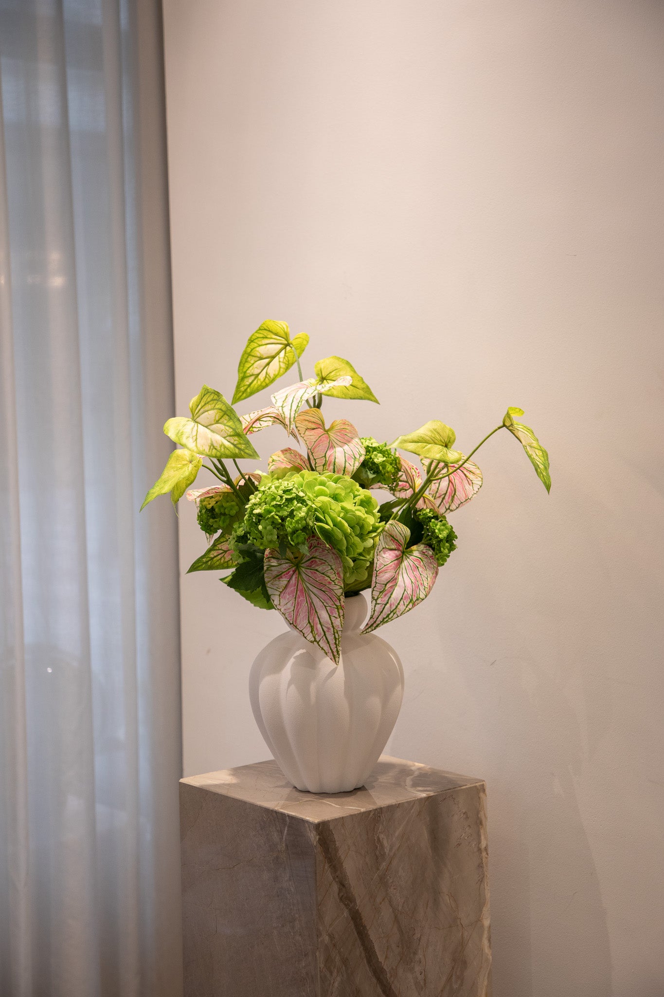 Billede af en kunstig blomster buket i grønne og lyserøde nuancer, placeret i en hvid keramik vase. Buketten står på en marmor piedestal.