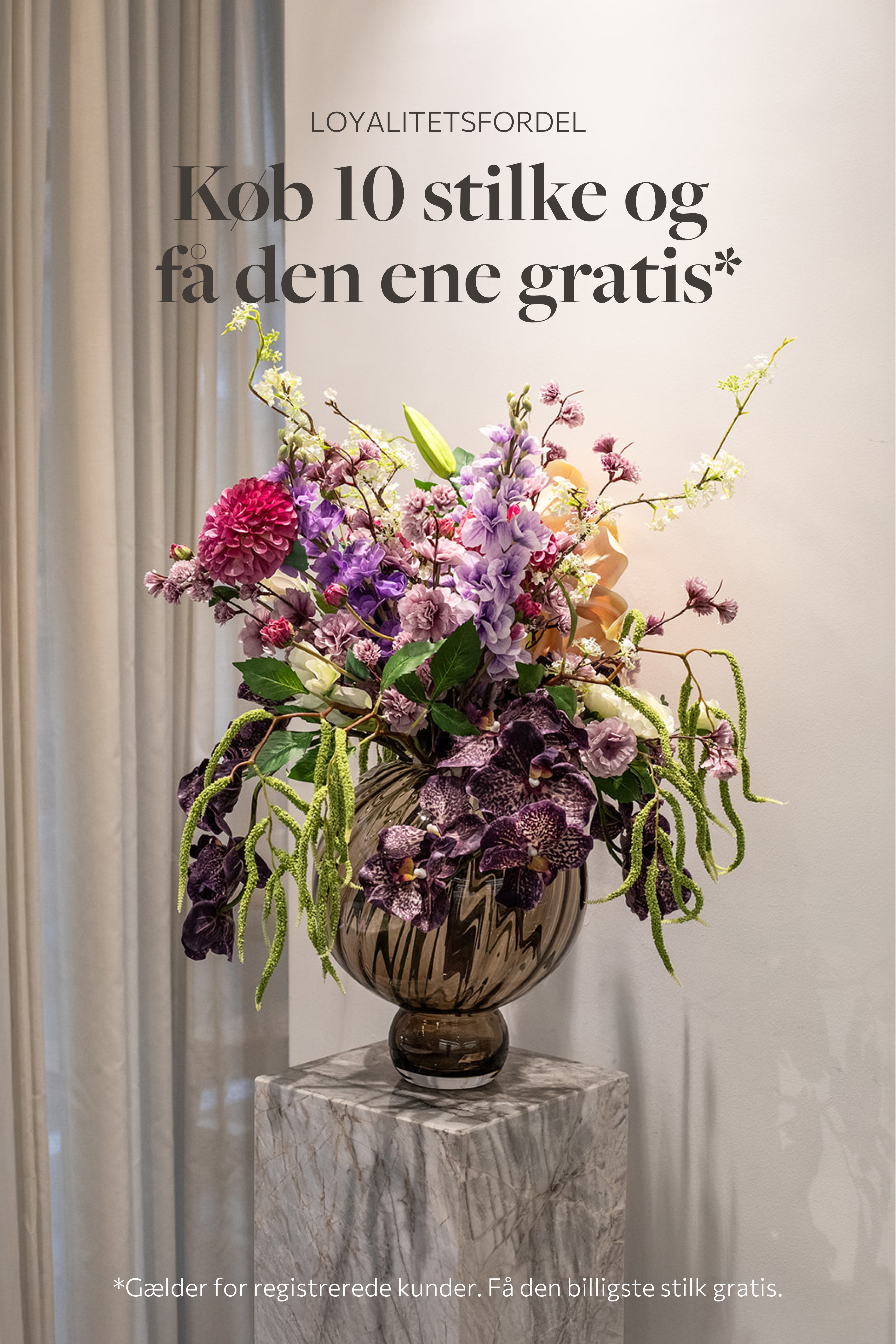 Stor farverig buket af kunstige blomster i masser af farver. Placeret i en stor topaz farvede glasvase, der står på en marmorpedestal.