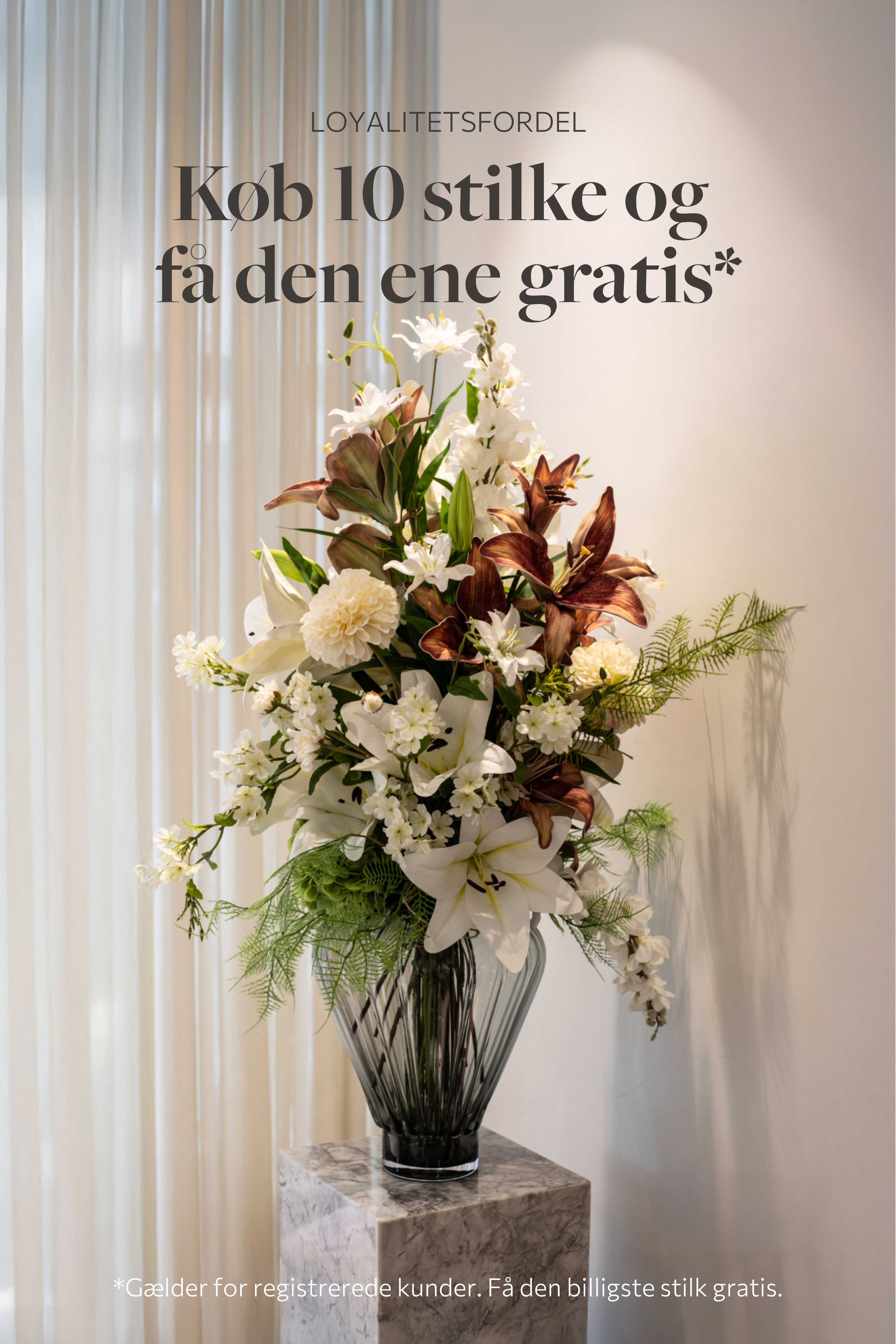 Stor farverig buket af kunstige blomster, placeret i en stor glasvase, der står på et marmor bord.