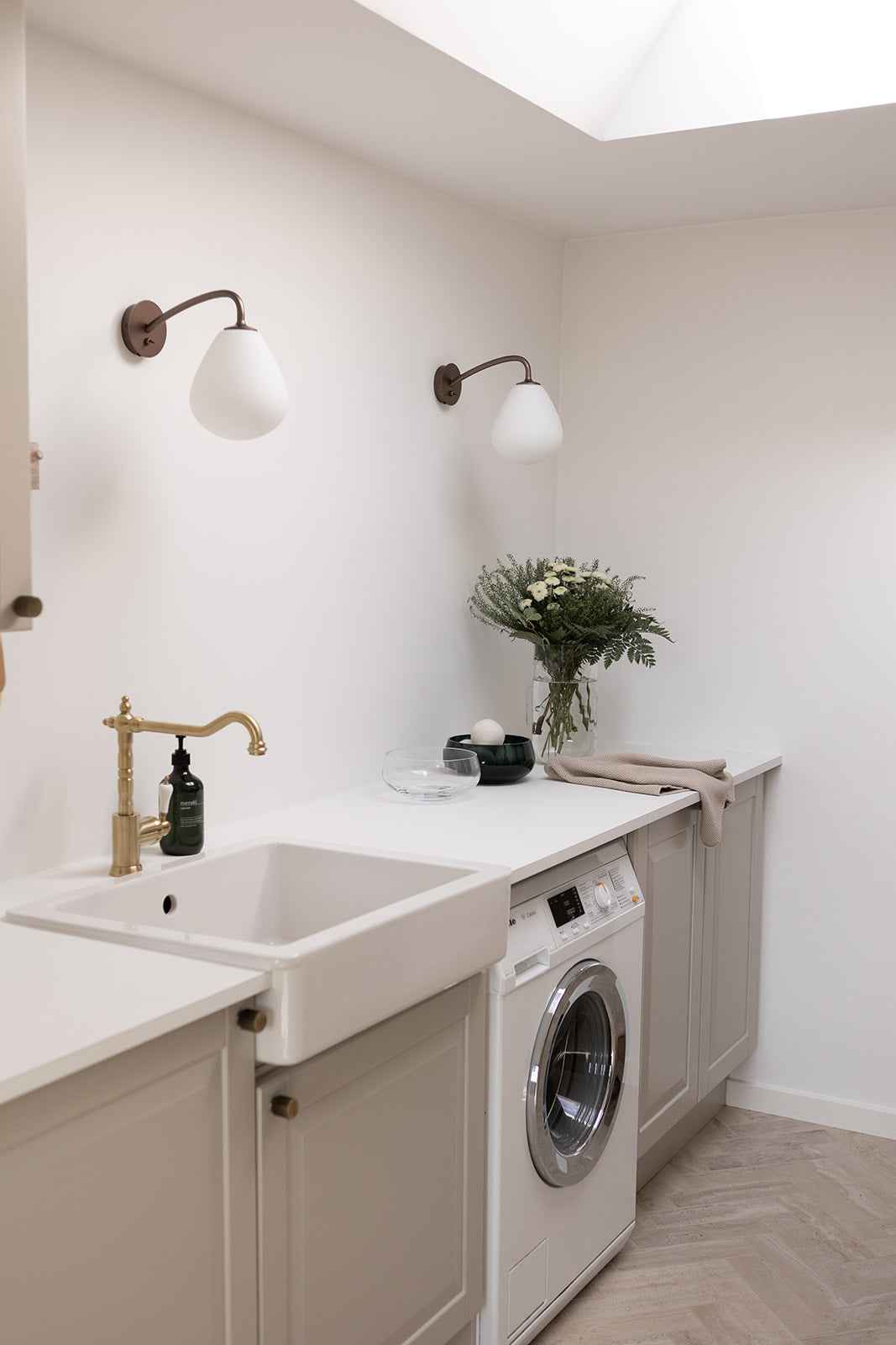 Et lyst vaskerum med hvide vægge og lysegrå skabe. På væggen hænger to væglamper med hvide glasskærme og mørke armaturer. Bordpladen rummer en indbygget vask med messingarmatur, en glasvase med blomster, en sort skål og et beige håndklæde. Under bordpladen står en frontbetjent vaskemaskine, og gulvet er lagt i sildebensmønster.