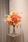 Kunstig buket i orange og hvide farver, der står i en gennemsigtig rund glas vase ovenpå en marmor piedestal.