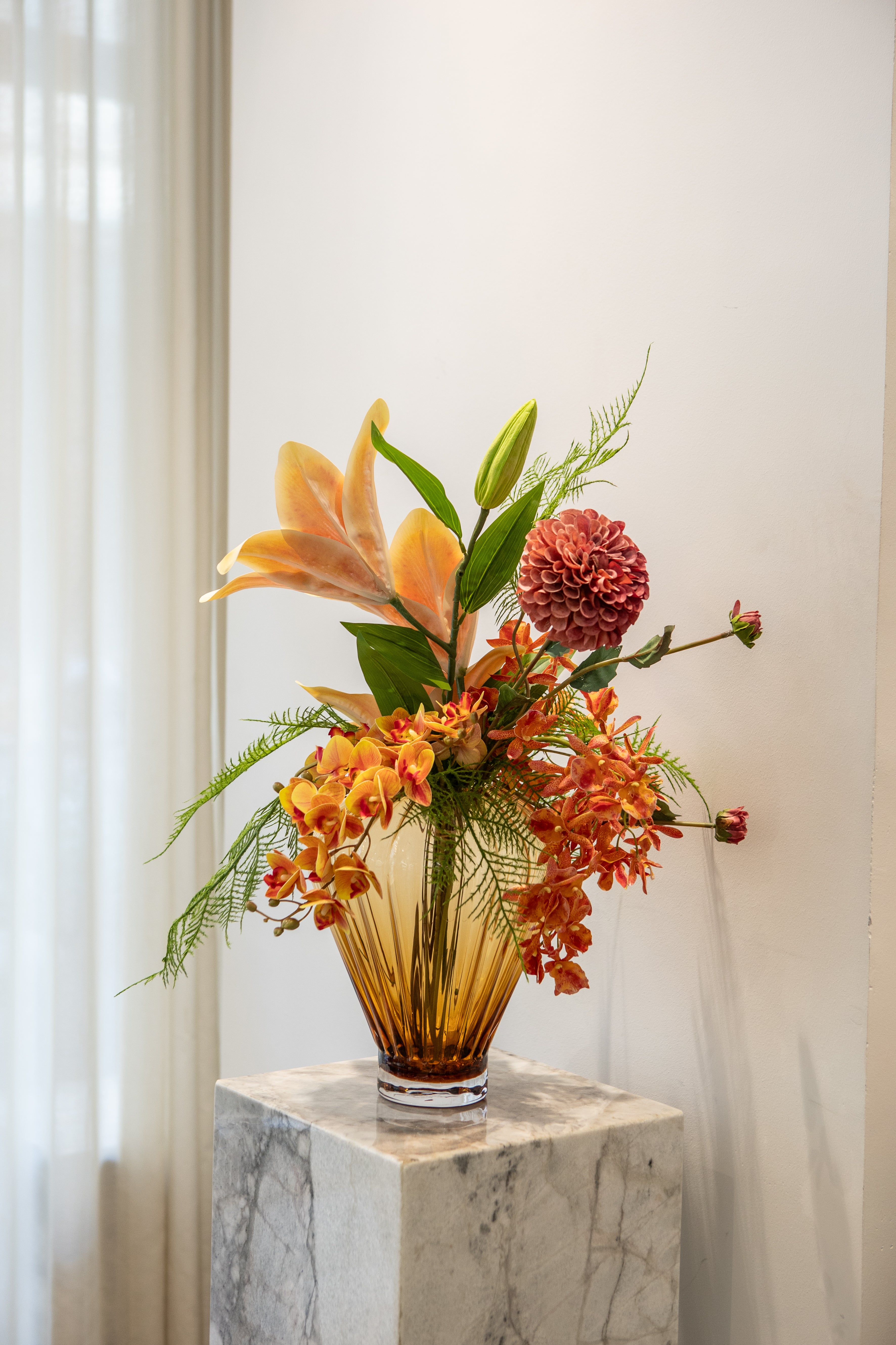 En stor buket af naturtro kunstige blomster  i orange og grønne nuancer i en stor amber farvet glasvase, der står på en marmorpedestal.