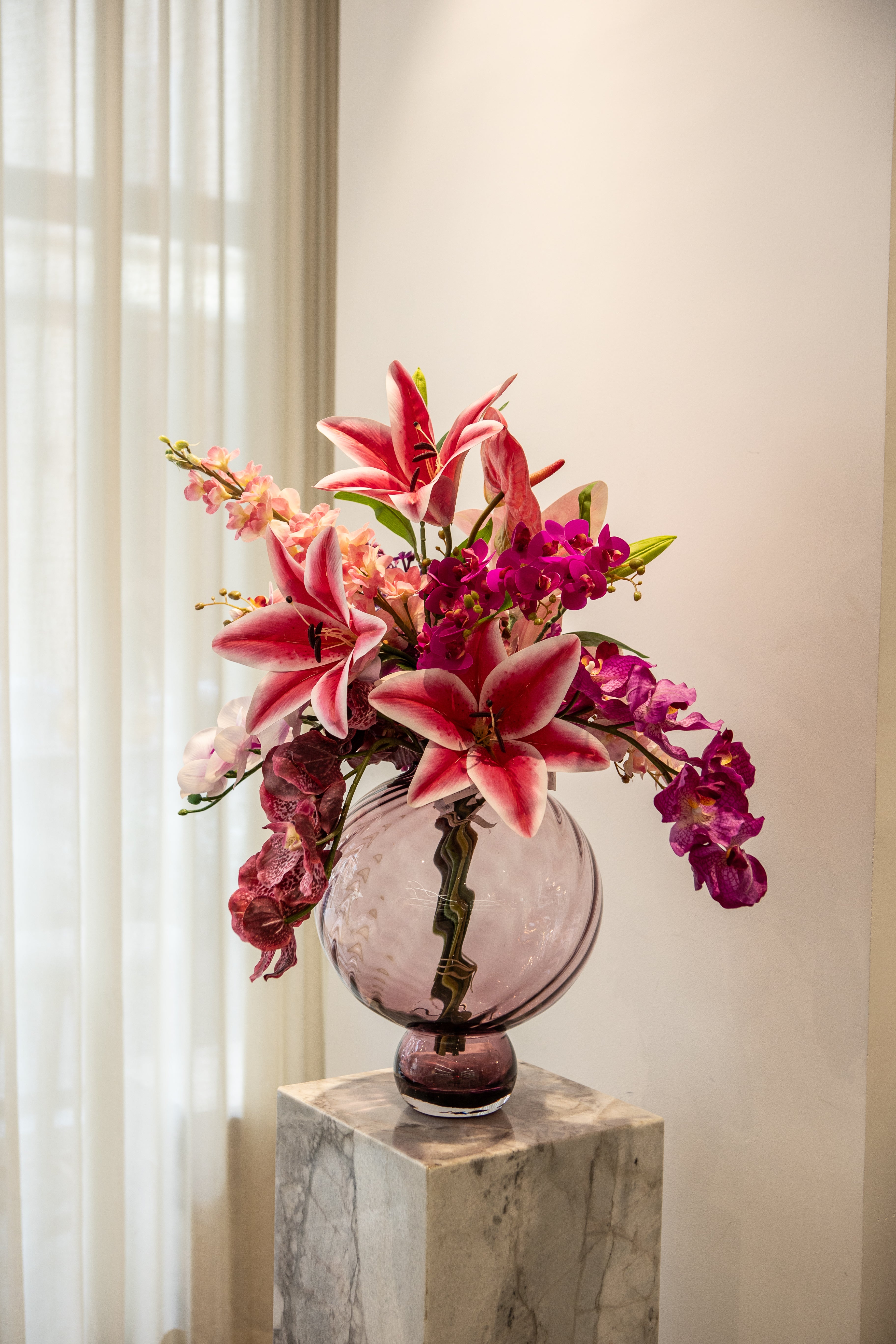 Stor buket af kunstige blomster i lilla og lyserøde farver i en plum farvet glasvase, der står på en marmorpedestal.