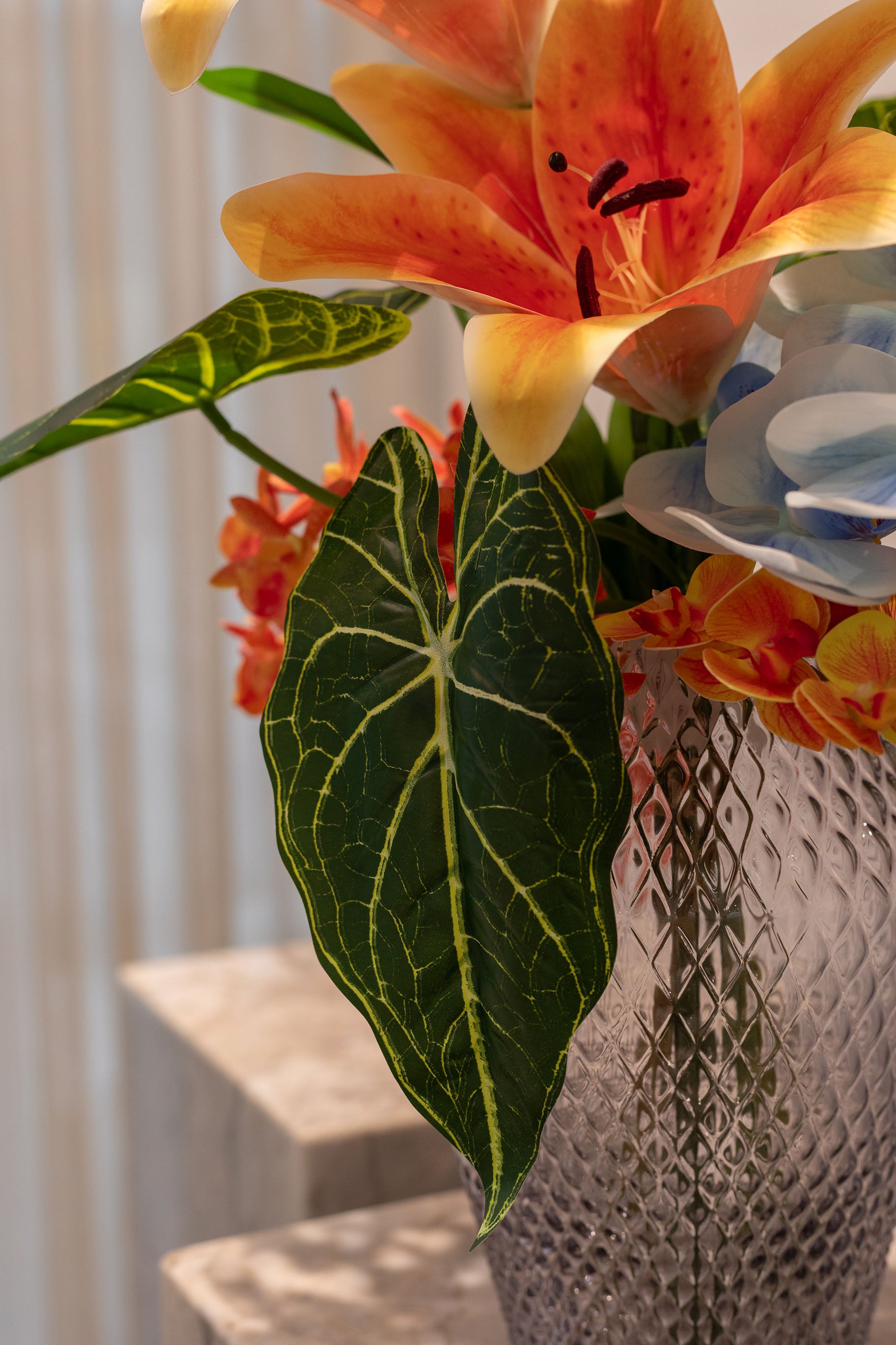 Nærbillede af buket af kunstige blomster i orange, blå og grønne farver. Placeret i en mønstret glasvase, der står på en marmor pedestal