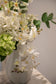 Nærbillede af buket af kunstige blomster i hvide og grønne nuancer. Placeret i en hvid keramikvase, der står på en marmor pedestal.