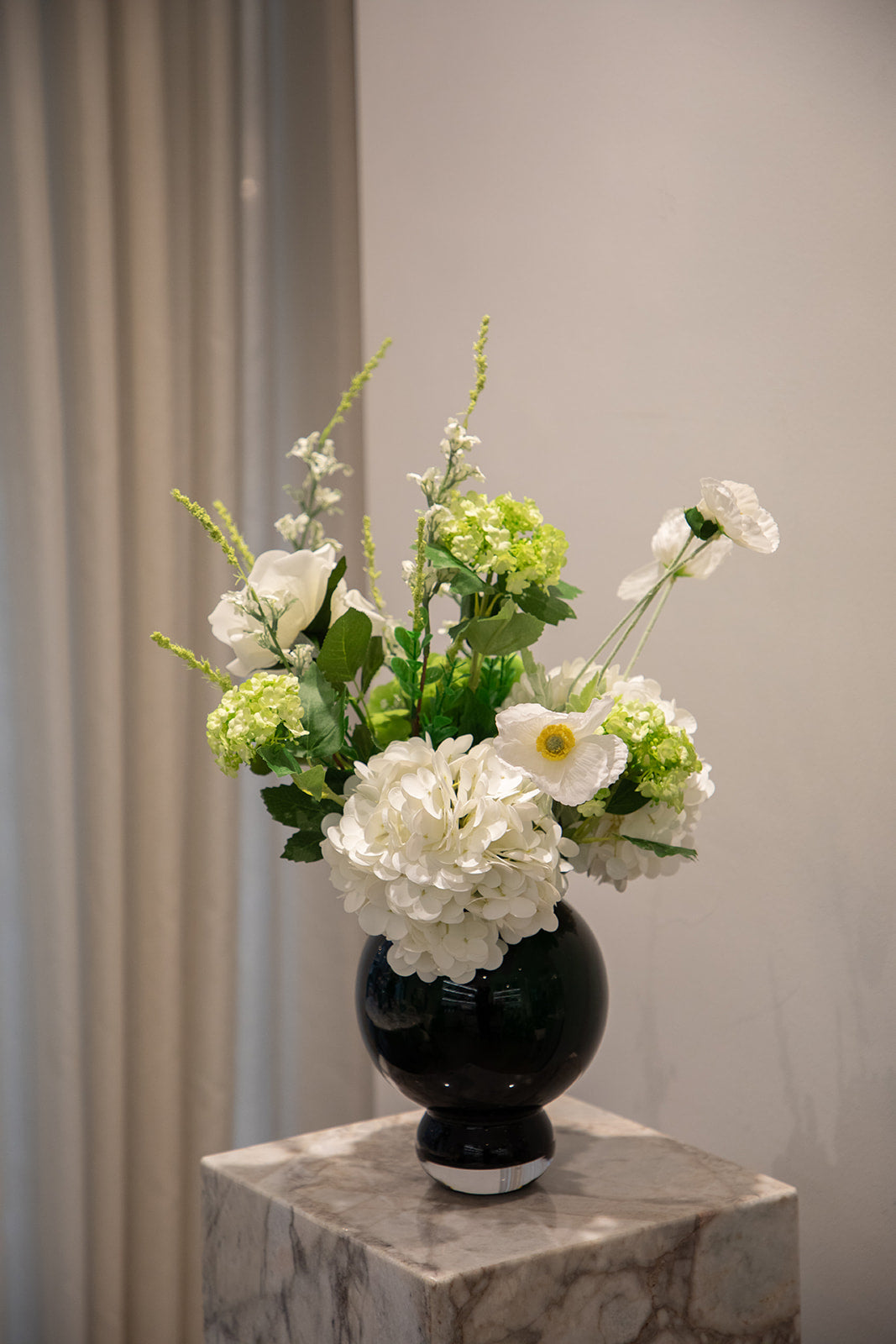 Buket af kunstige blomster i grønne og hvide nuancer i en blå glasvase. Står på en marmor pedestal.