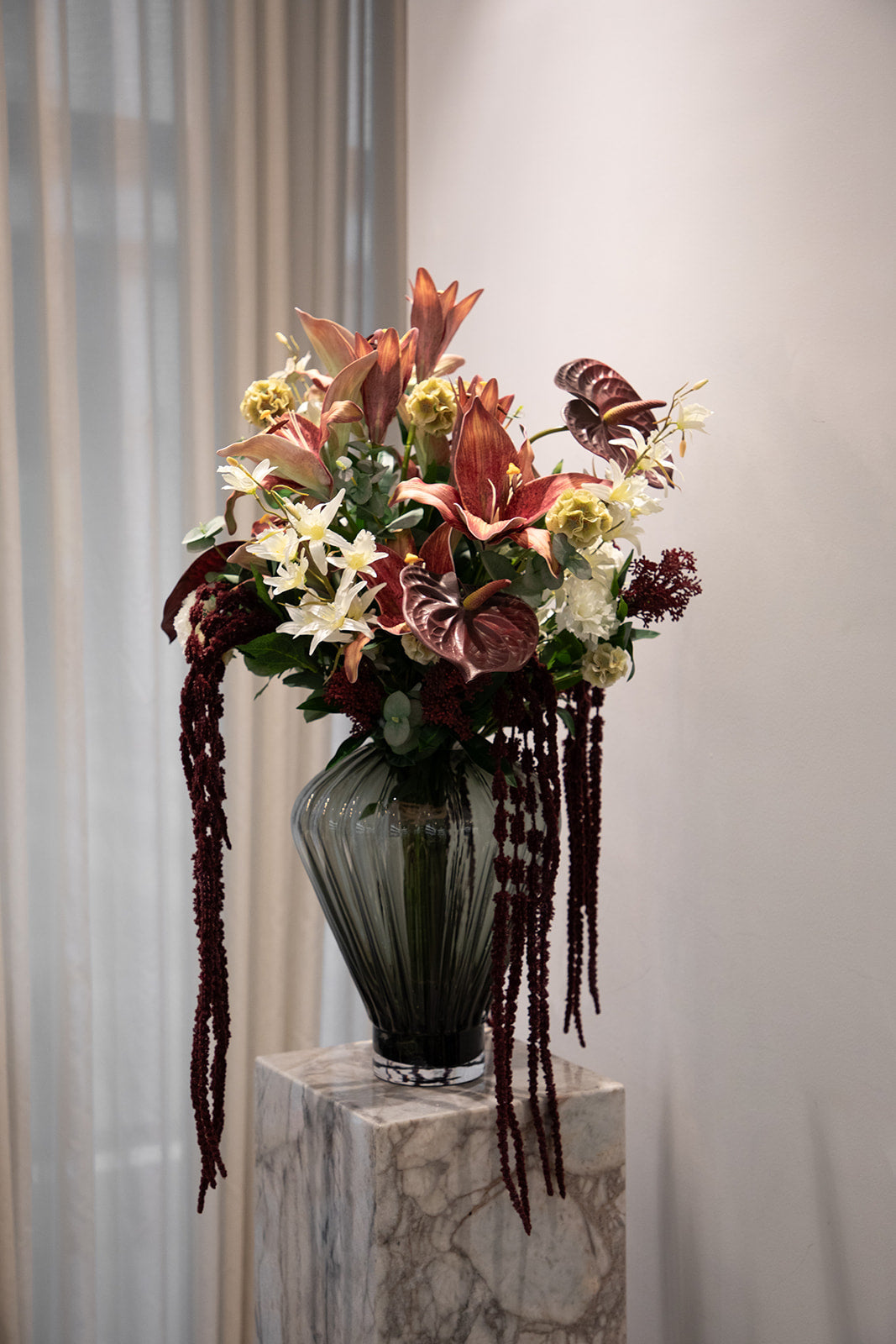 Stor buket af kunstige blomster i røde, hvide og creme nuancer. Placeret i en grå glasvase, der står oven på en marmor pedestal.