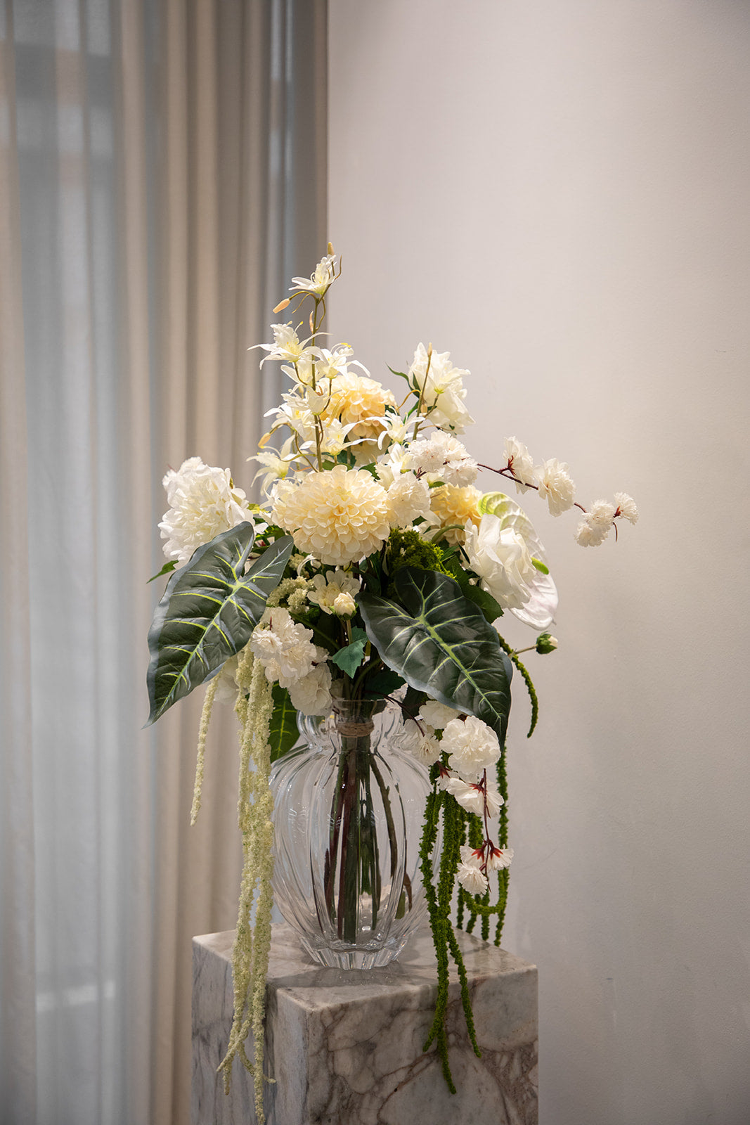 Stor buket af kunstige blomster i grønne og hvide nuancer. Placeret i en gennemsigtig glasvase, der står oven på en marmor pedestal.