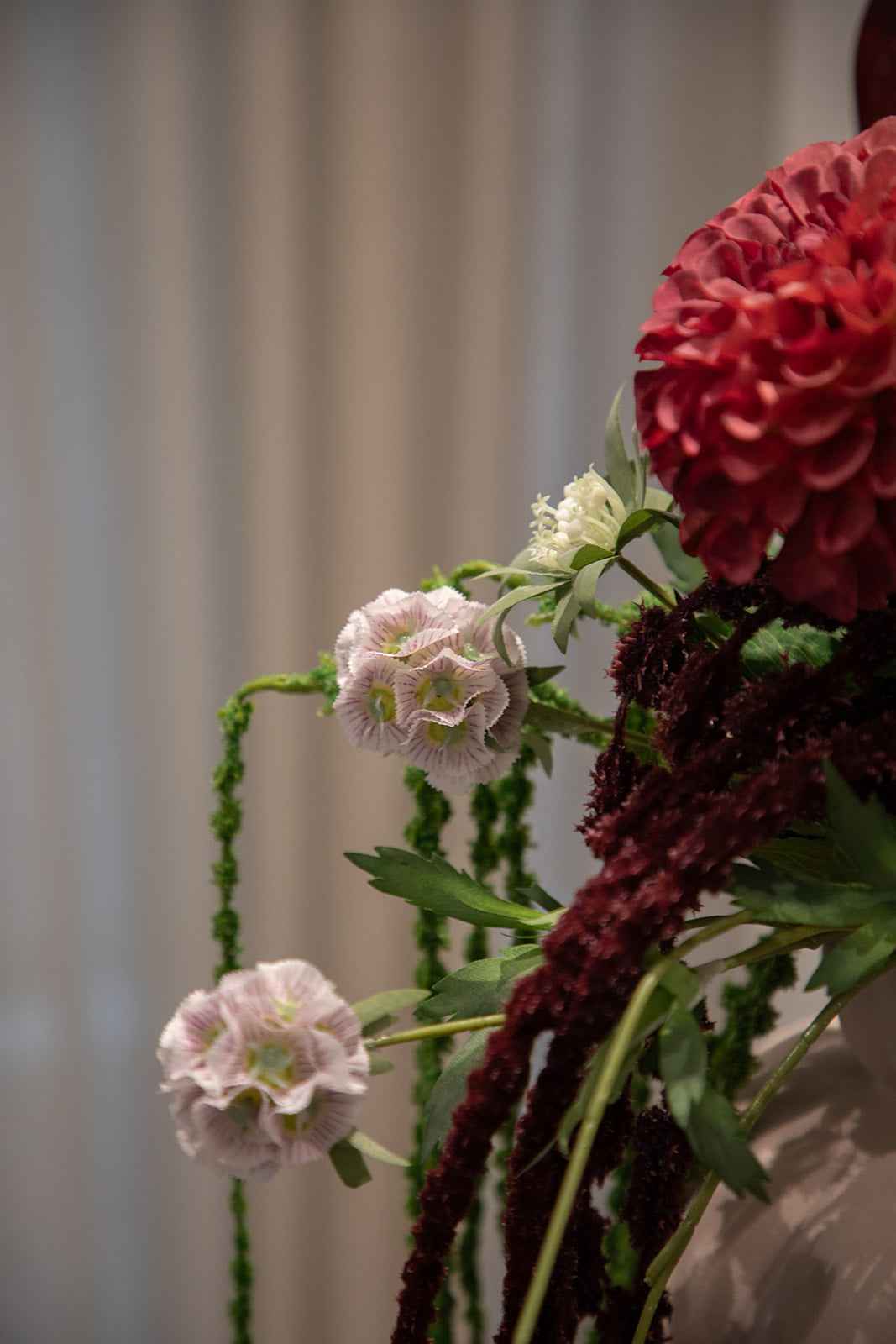 Nærbillede af buket af kunstige blomster i røde, grønne og rosa nuancer. Placeret i en sand farvede keramikvase.