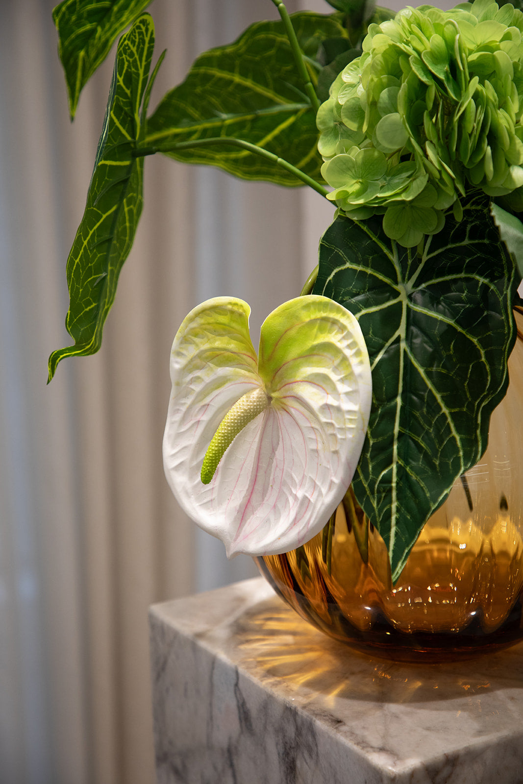 Nærbillede af kunstig buket i grønne nuancer. Buketten står i en orange farvet glasvase, der står oven på en marmor pedestal.