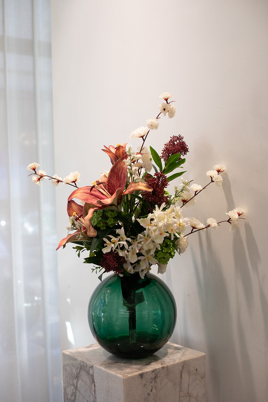 Kunstig buket i hvide, grønne og røde nuancer. placeret i en grøn glas vase, der står på en marmor pedestal.