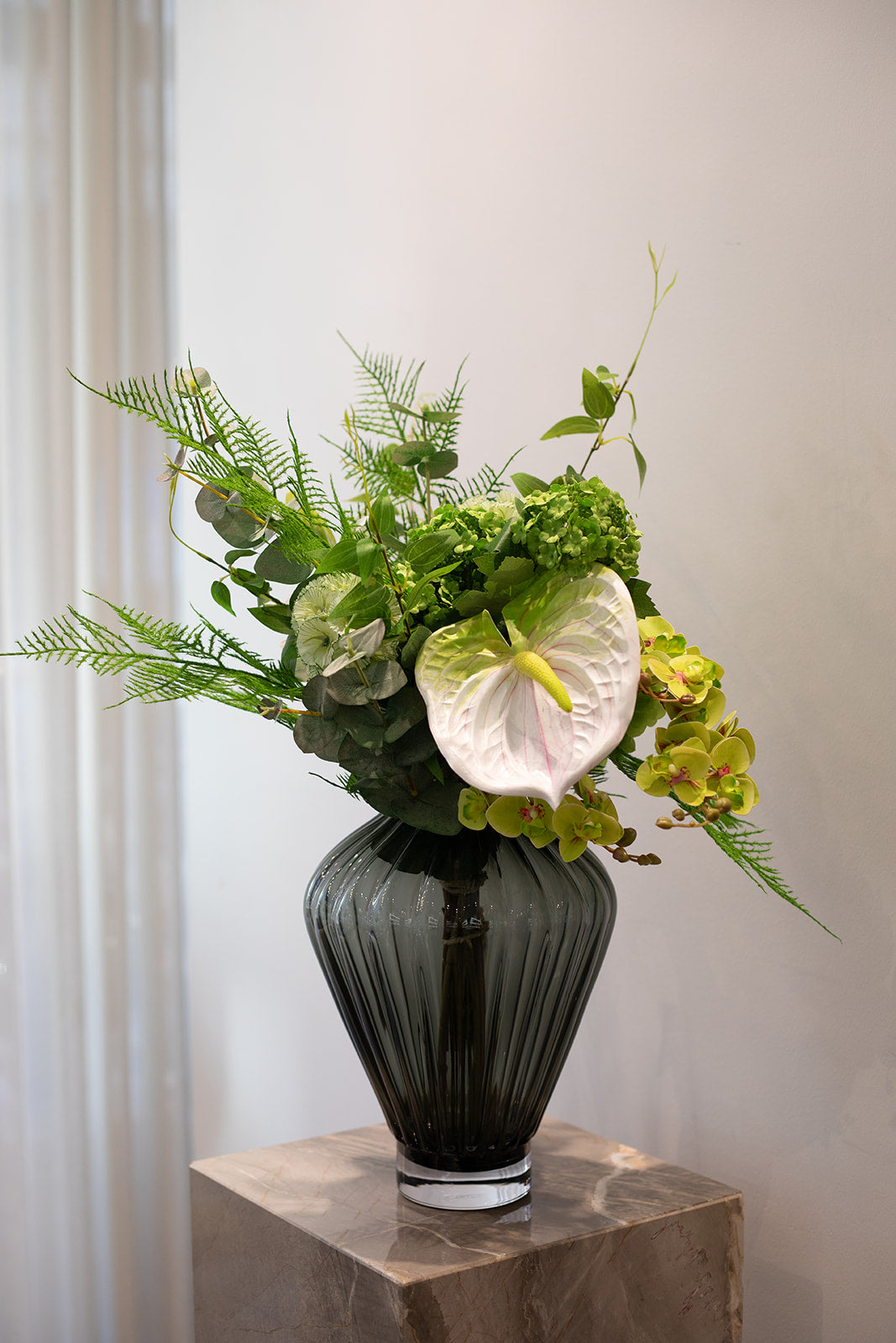 Buket af kunstige blomster i grønne og hvide toner. Buketten er placeret i en mørk grå glas vase, der står ovenpå en marmor pedestal.