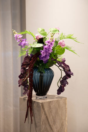 Billede af kunstig buket i røde, lilla og grønne nuancer. Buketten står en i en blå glas vase oven på en marmor piedestal.