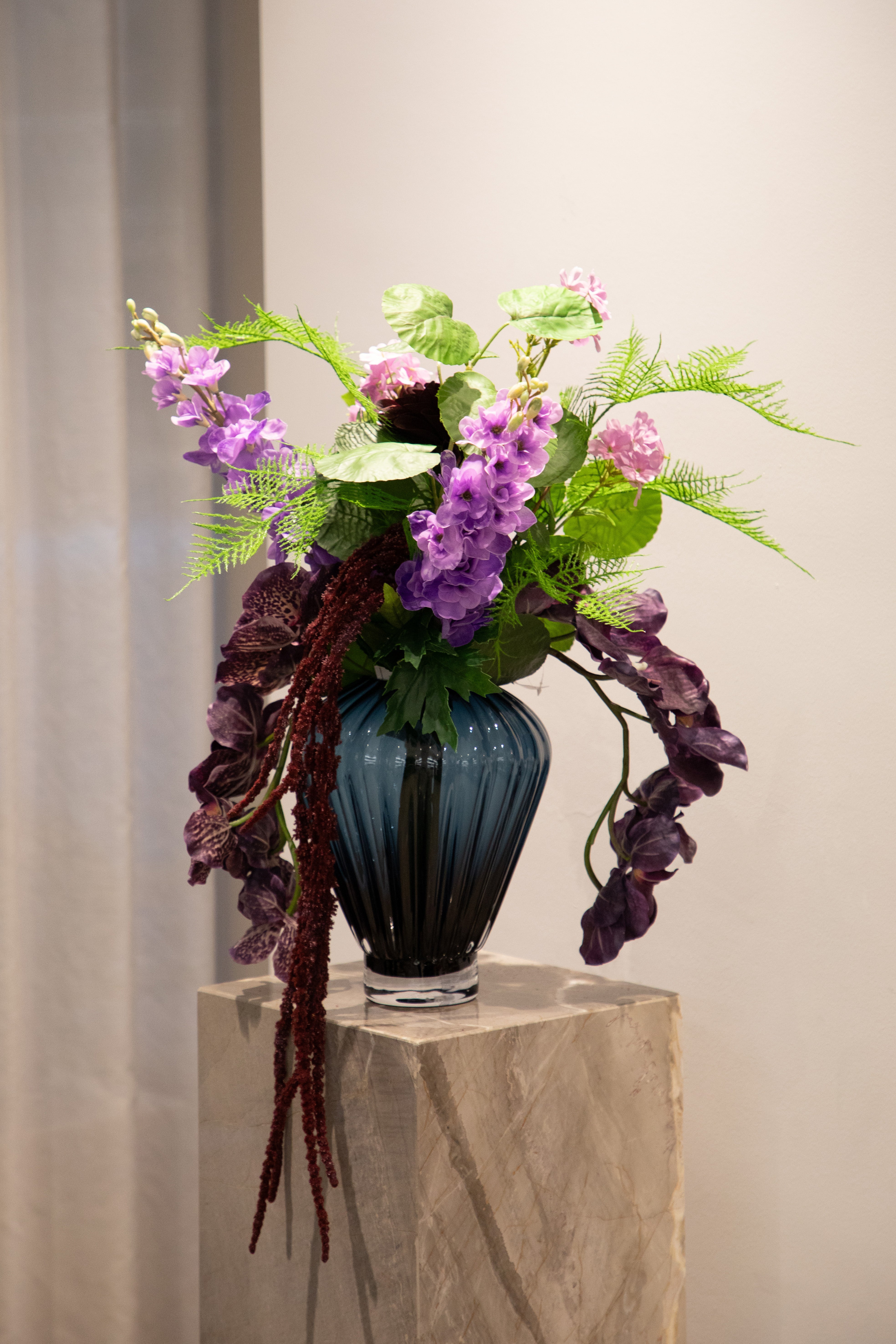 Billede af kunstig buket i røde, lilla og grønne nuancer. Buketten står en i en blå glas vase oven på en marmor piedestal.