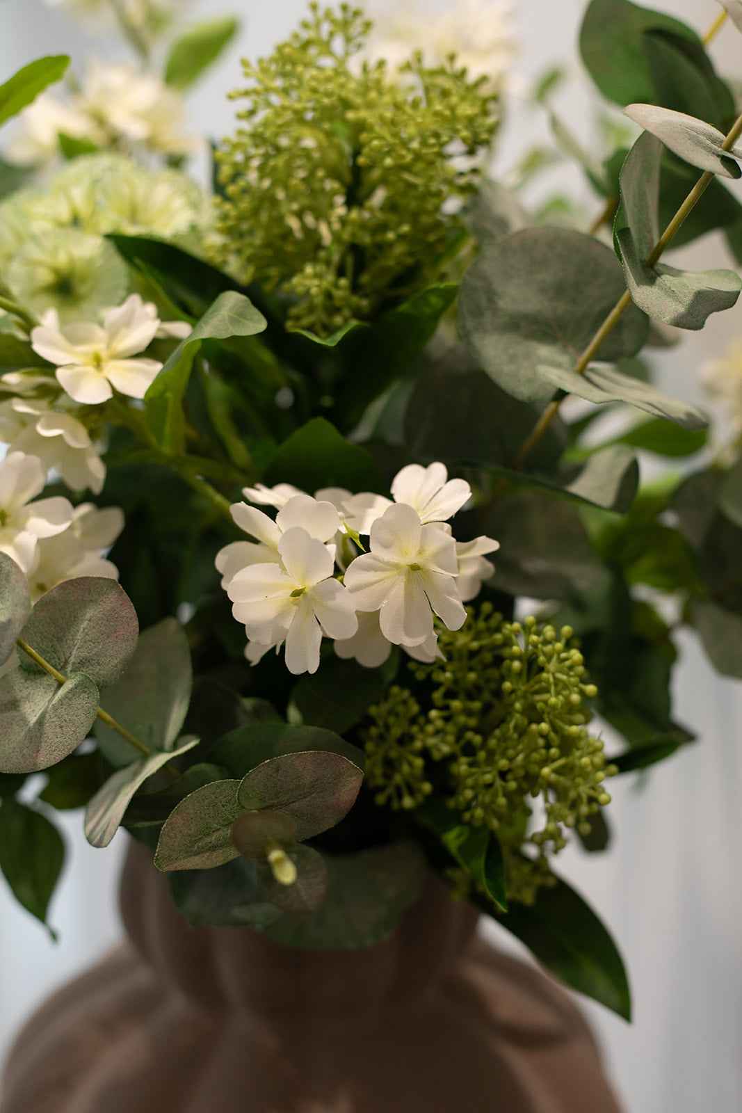 Nærbillede af kunstig buket i grønne og hvide nuancer, placeret i en brun keramik vase.