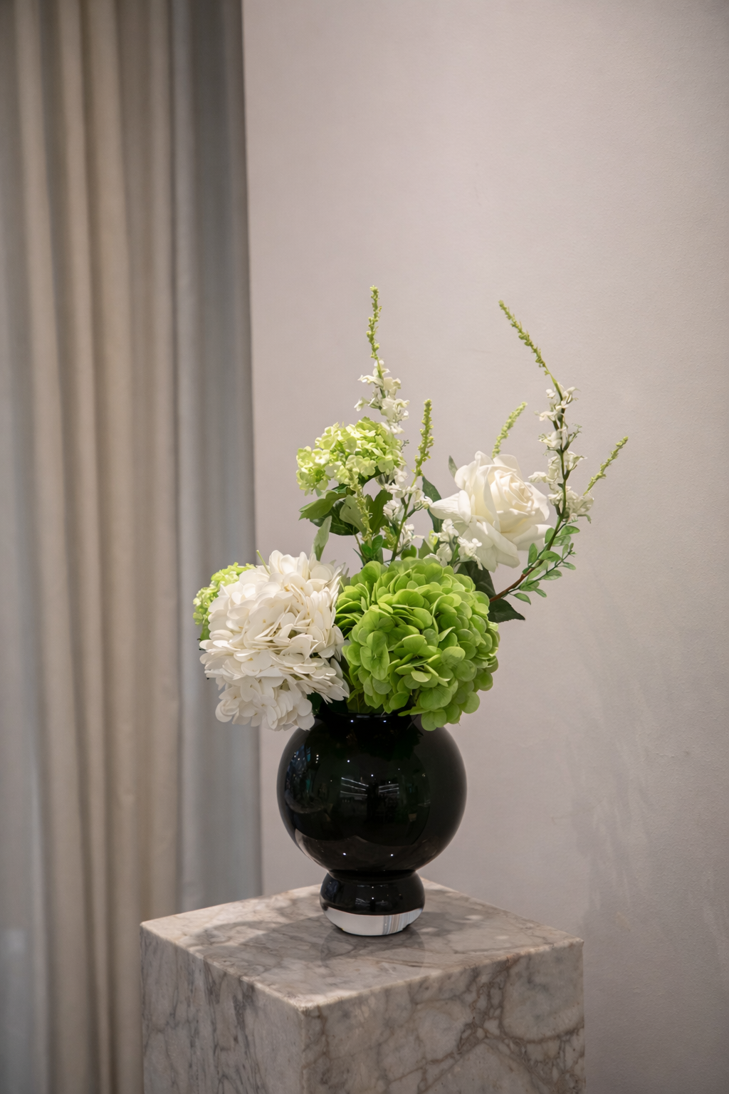 Buket af kunstige blomster i grønne og hvide nuancer. Placeret i en lille blå glasvase, der står på en marmor pedestal.