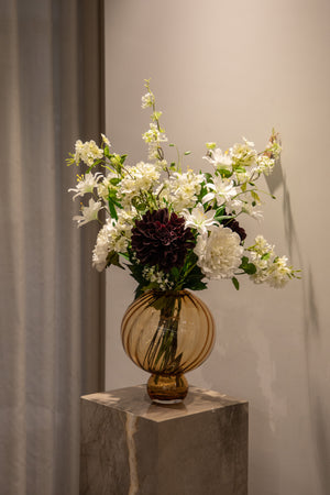 Kunstig buket i hvide og bordeaux farver, der står i en orange glasvase, ovenpå en marmor piedestal.