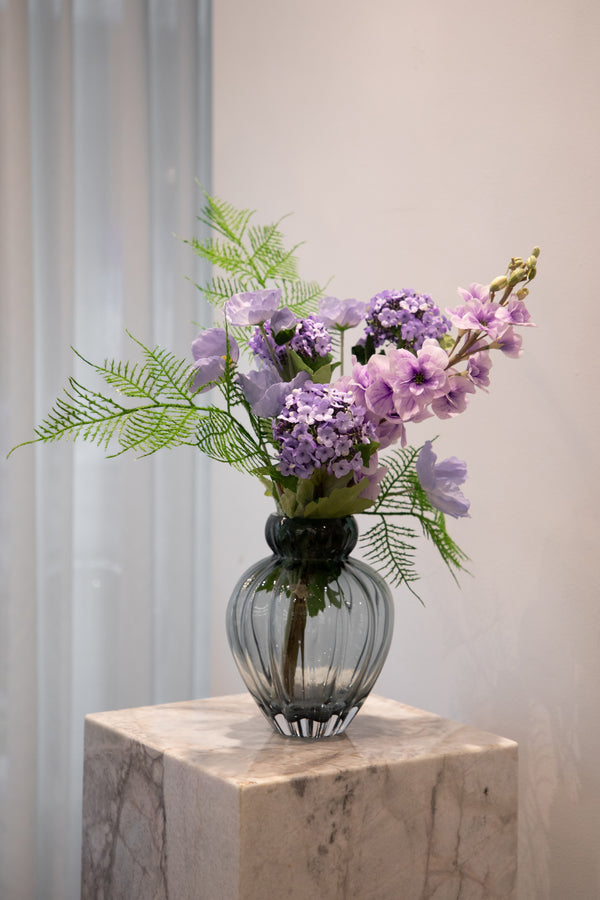 Kunstig buket i lilla og grønne nuancer, placeret i en grå glas vase, der står på en marmor pedestal.