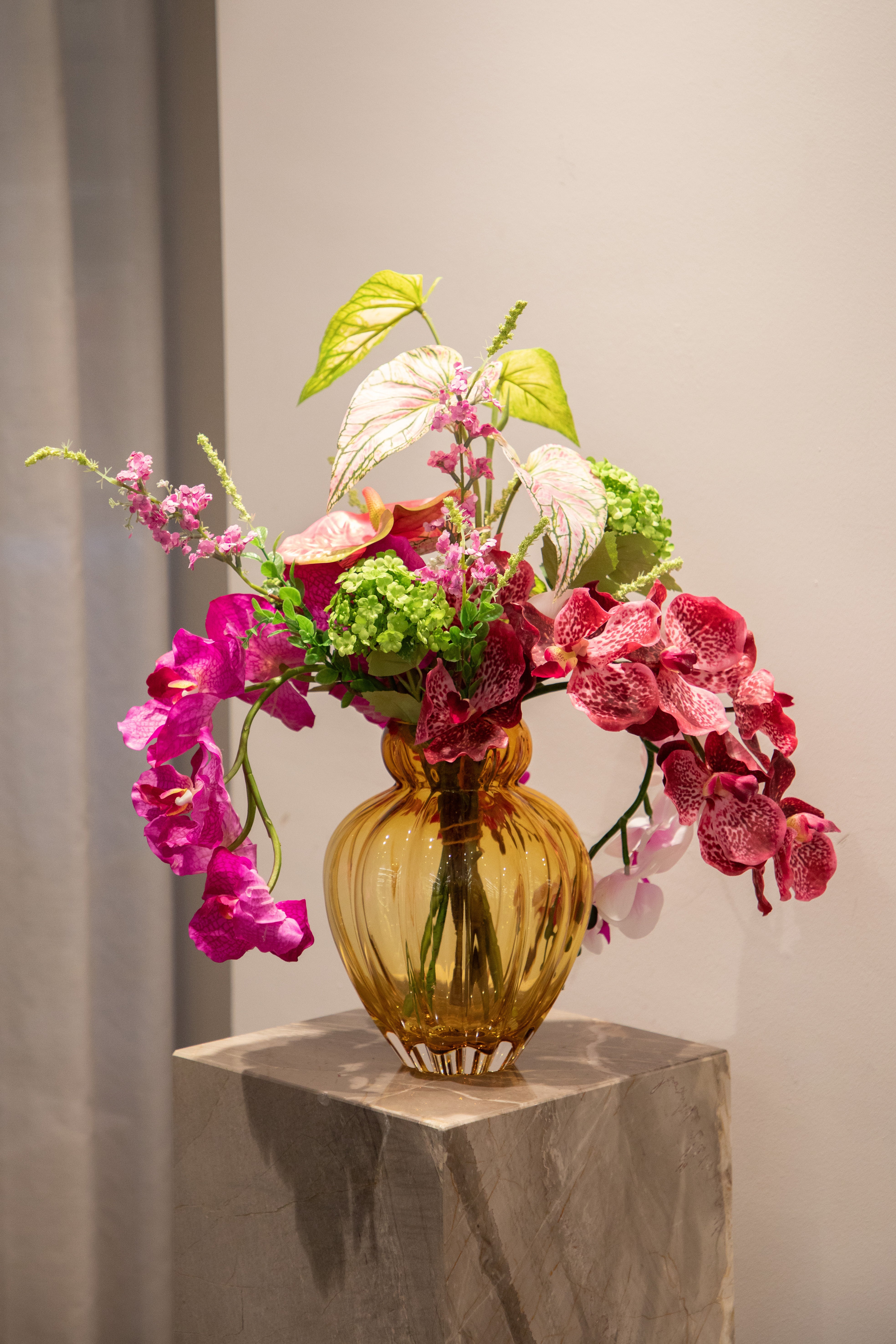 Kunstig buket blomster i lyserøde, grønne og rosa nuancer. Placeret i en orange glas vase, der står på en marmor piedestal.
