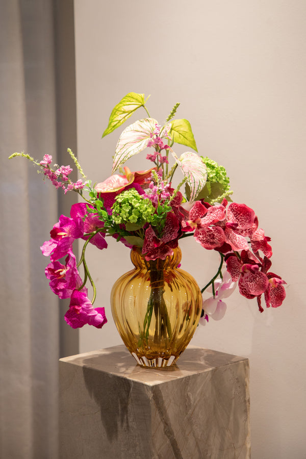Kunstig buket blomster i lyserøde, grønne og rosa nuancer. Placeret i en orange glas vase, der står på en marmor piedestal.