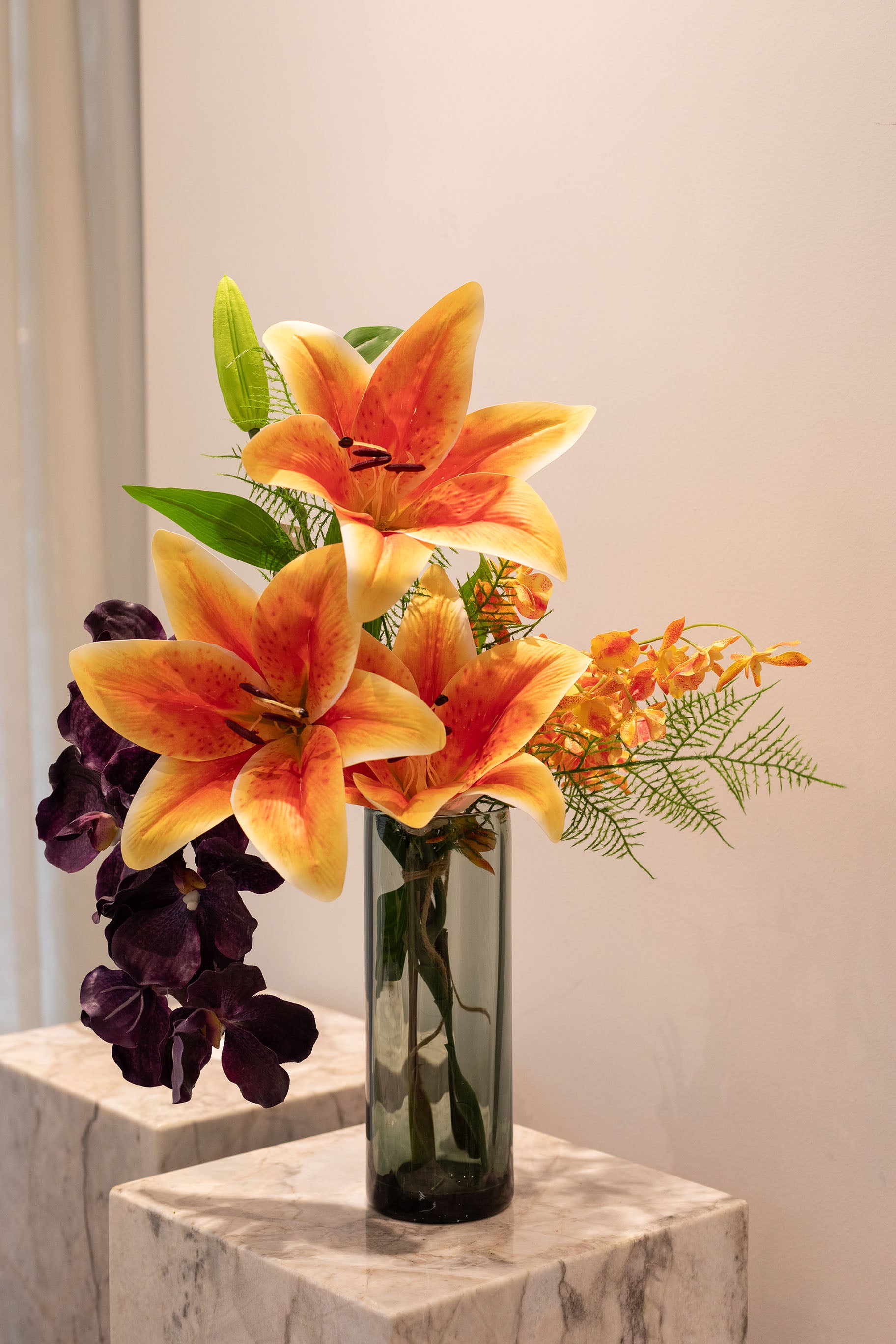 Buket af kunstige blomster i orange, mørke lilla og grønne nuancer. Placeret i en røgfarvede glasvase, der står på en marmor pedestal.