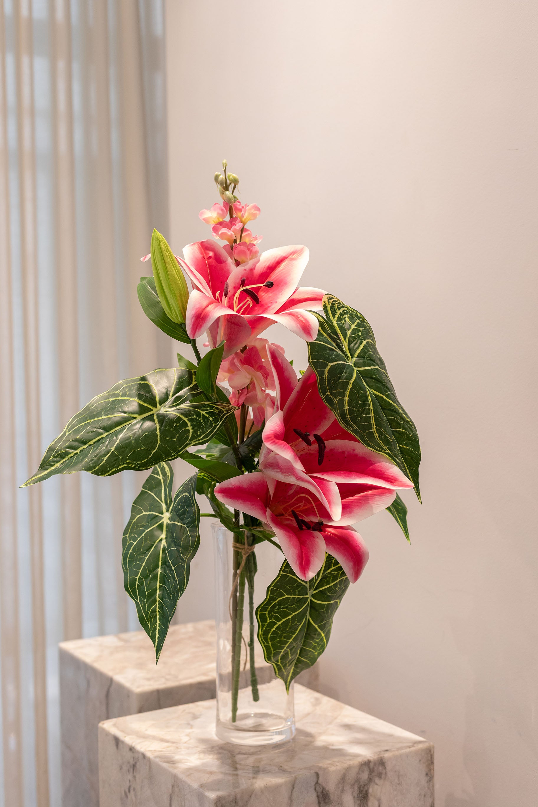 Buket af kunstige blomster i lyserøde og grønne nuancer. Placeret i en gennemsigtig glasvase, der står på en marmor pedestal.