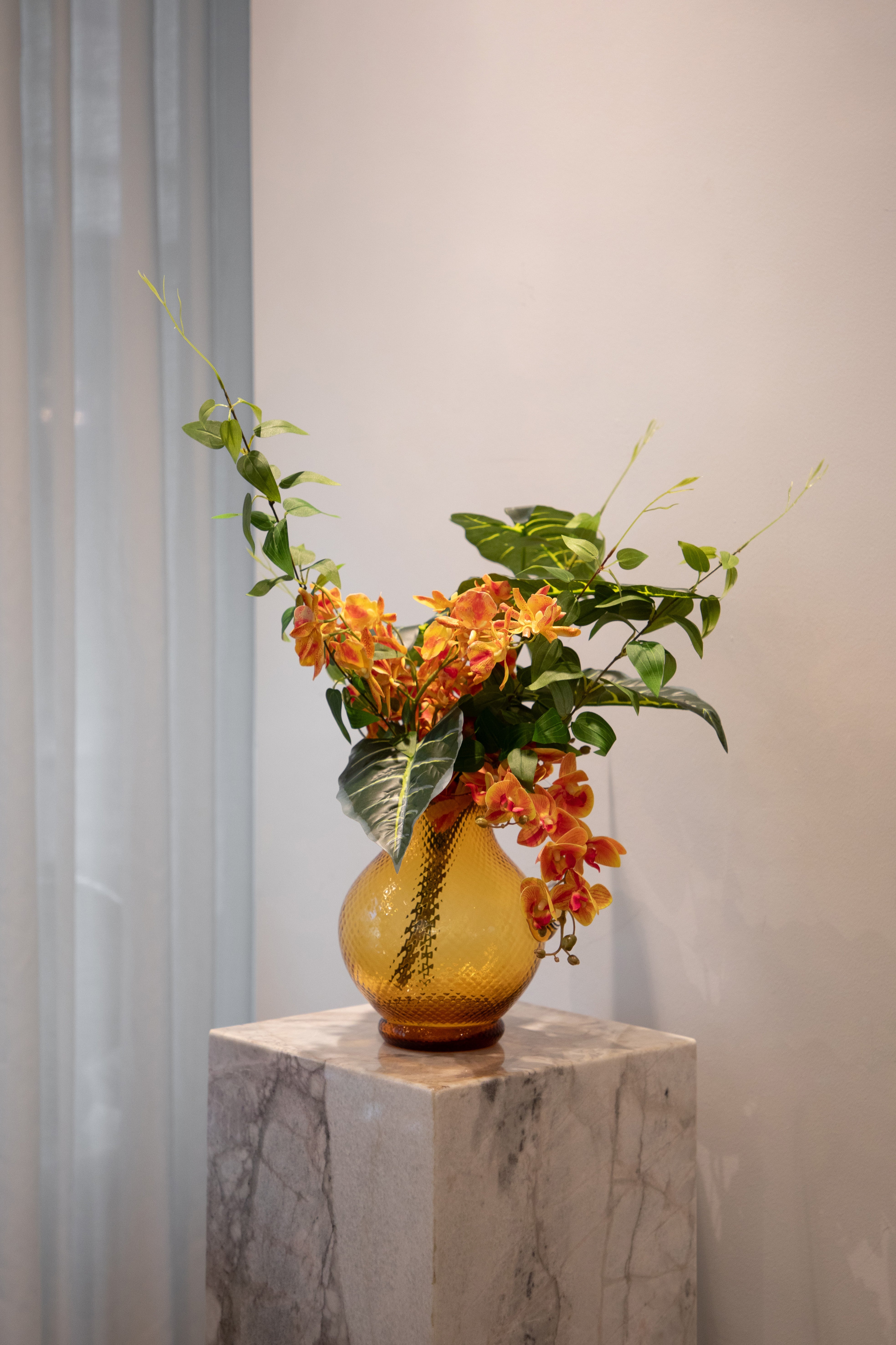 Kunstig buket i grønne og orange nuancer, placeret i en orange glas kande, der står på en marmor pedestal.