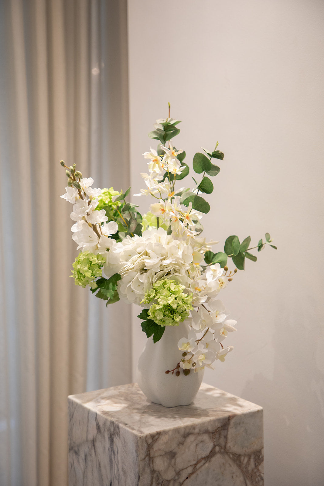 Buket af kunstige blomster i hvide og grønne nuancer. Placeret i en hvid keramikvase, der står på en marmor pedestal.