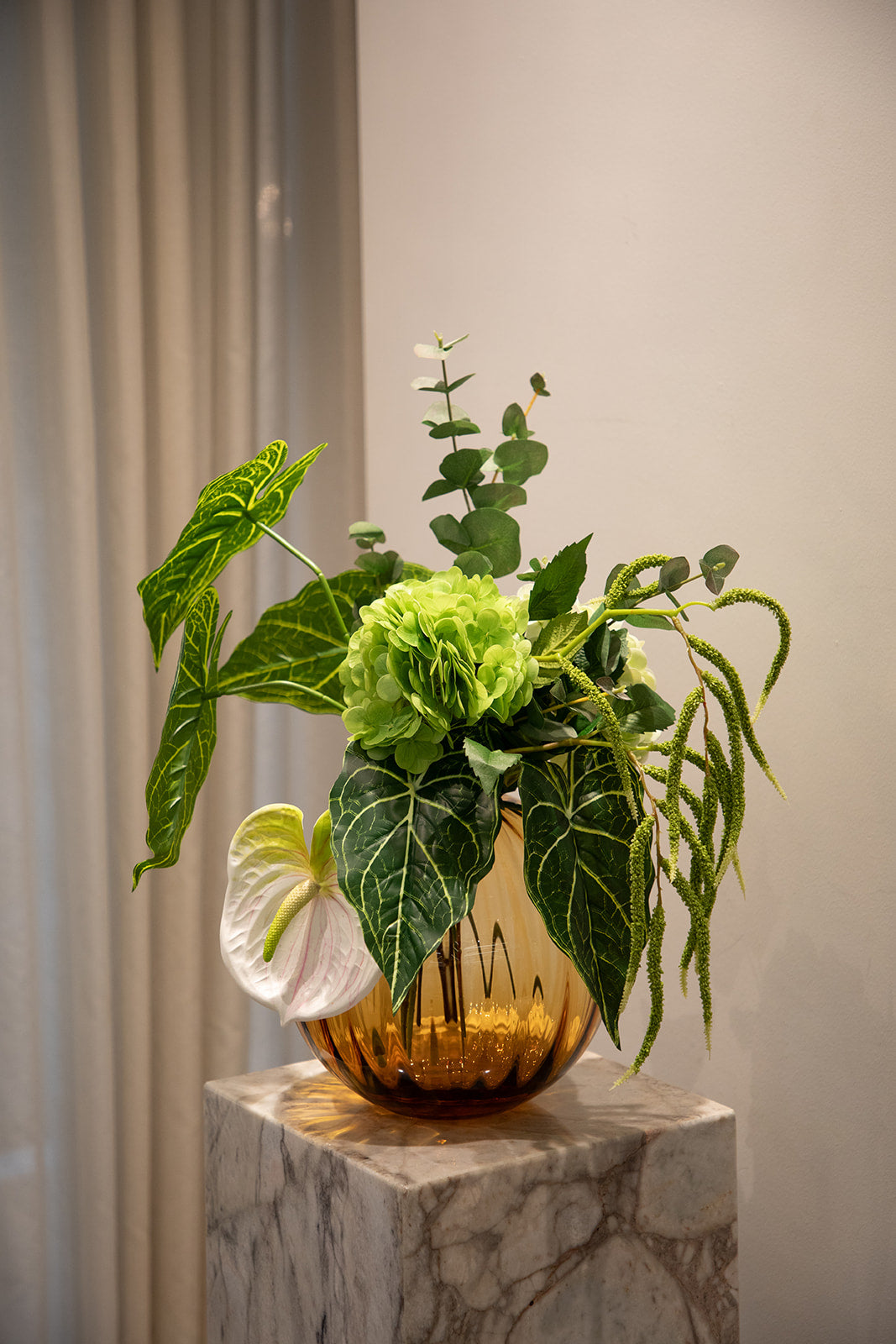 Stor buket i grønne nuancer. Placeret i orange farvet glasvase, der står oven på en pedestal.